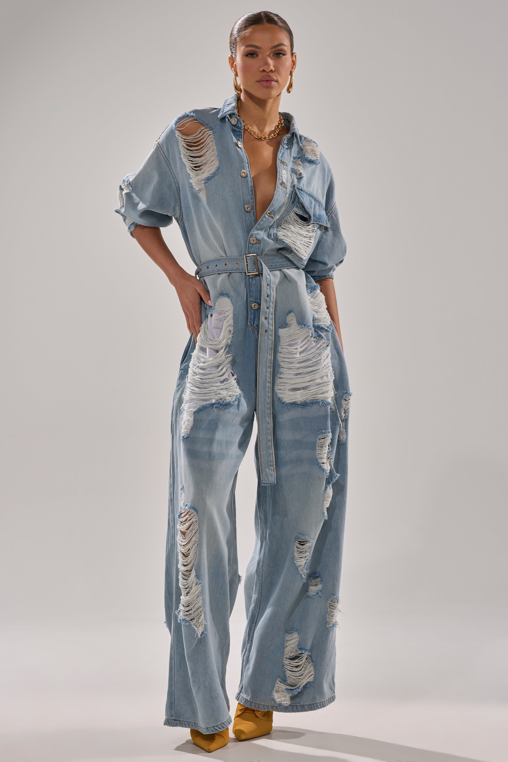 A woman confidently wears the NOT TODAY EXTREME OVERSIZED DISTRESSED DENIM JUMPSUIT featuring a belted waist, rolled-up sleeves, and wide legs, styled with gold jewelry and yellow heels against a plain gray background.