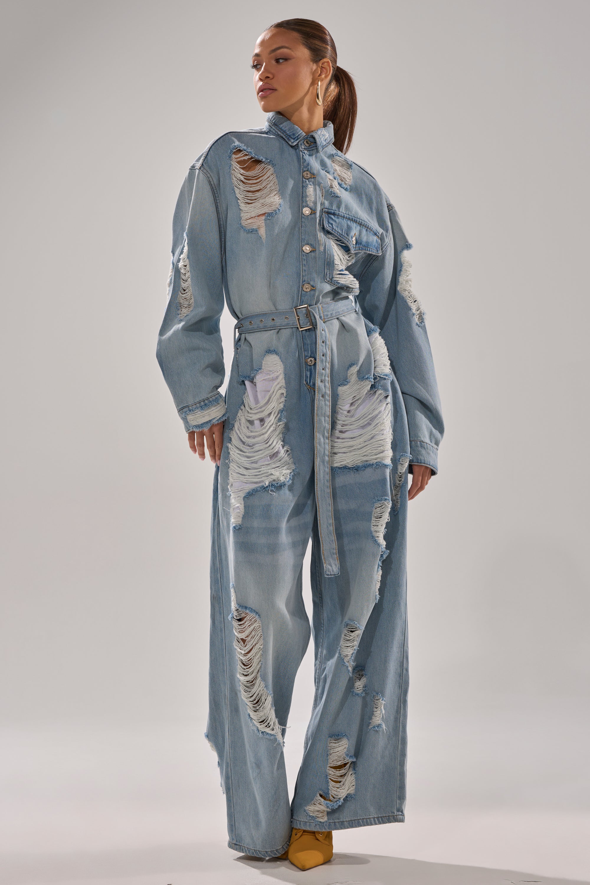A woman poses in the NOT TODAY EXTREME OVERSIZED DISTRESSED DENIM JUMPSUIT, featuring wide legs, a button front, and a matching belt. She completes her look with yellow pointed shoes and a sleek ponytail.