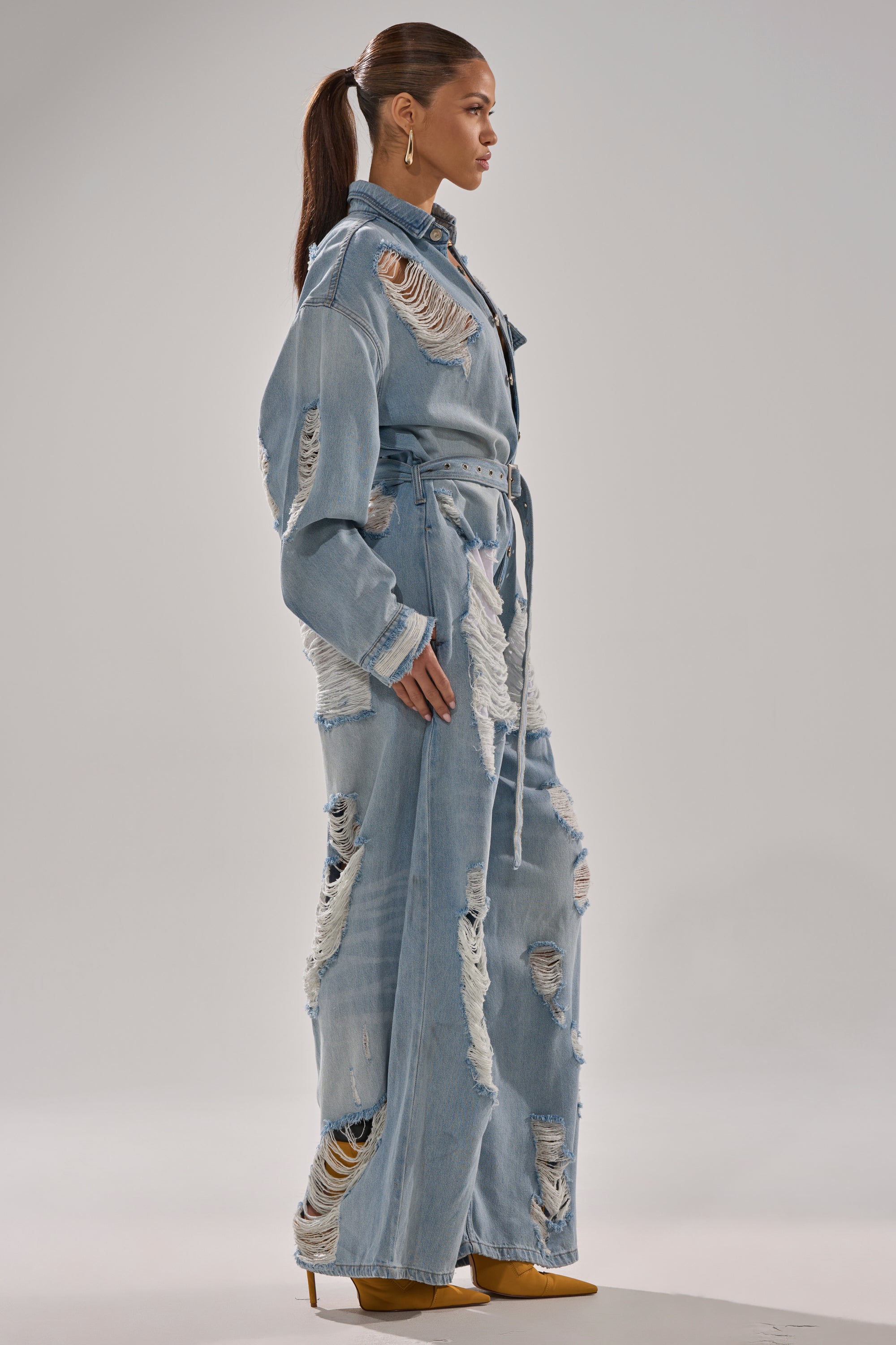 A woman in profile wears the NOT TODAY EXTREME OVERSIZED DISTRESSED DENIM JUMPSUIT with a wide-leg fit and adjustable waist belt, paired with tan heels. Her hair is styled in a sleek ponytail against a plain light gray background.