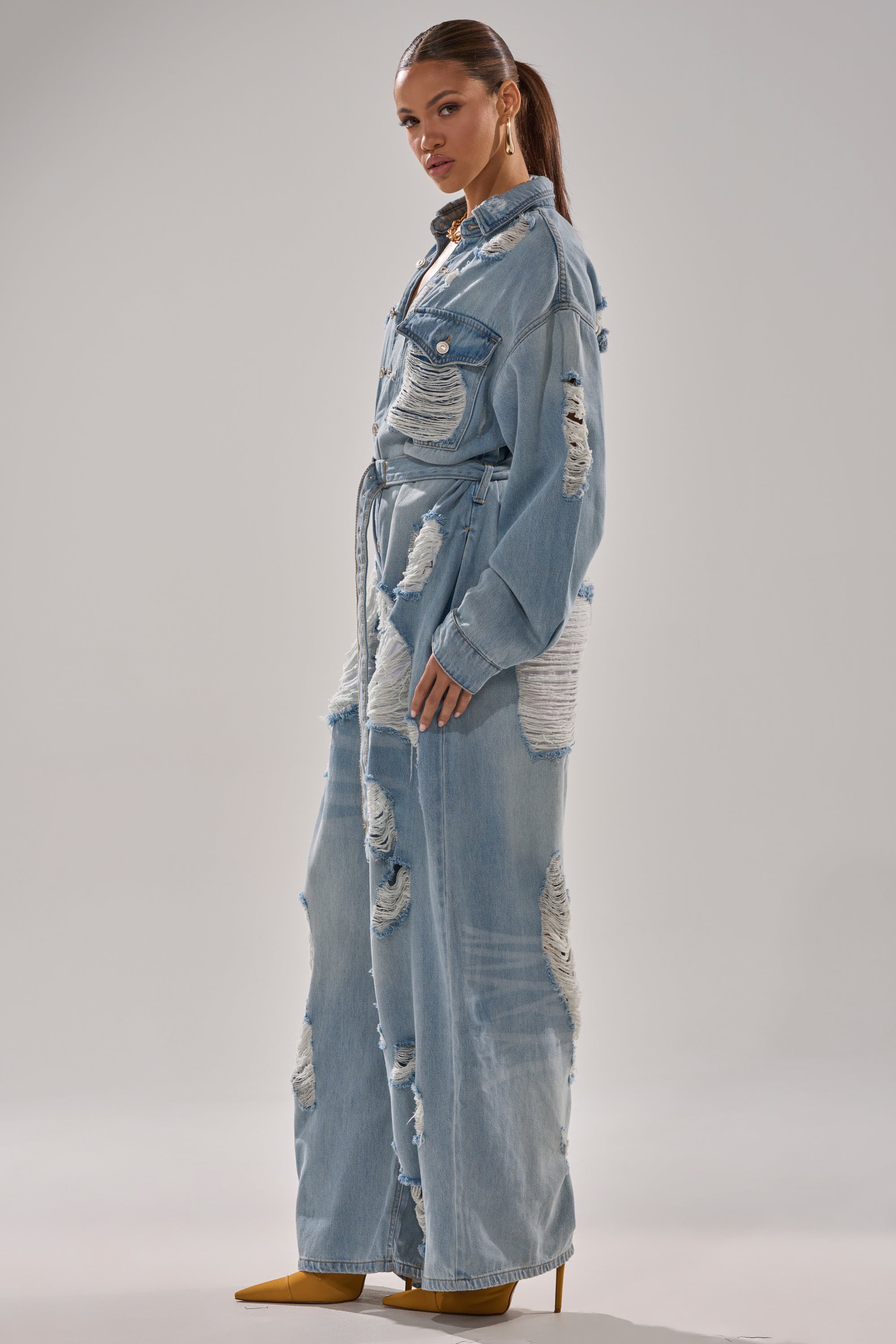 A woman in profile wears the NOT TODAY EXTREME OVERSIZED DISTRESSED DENIM JUMPSUIT with chest pockets and wide legs, paired with yellow pointed-toe heels, against a plain light background.