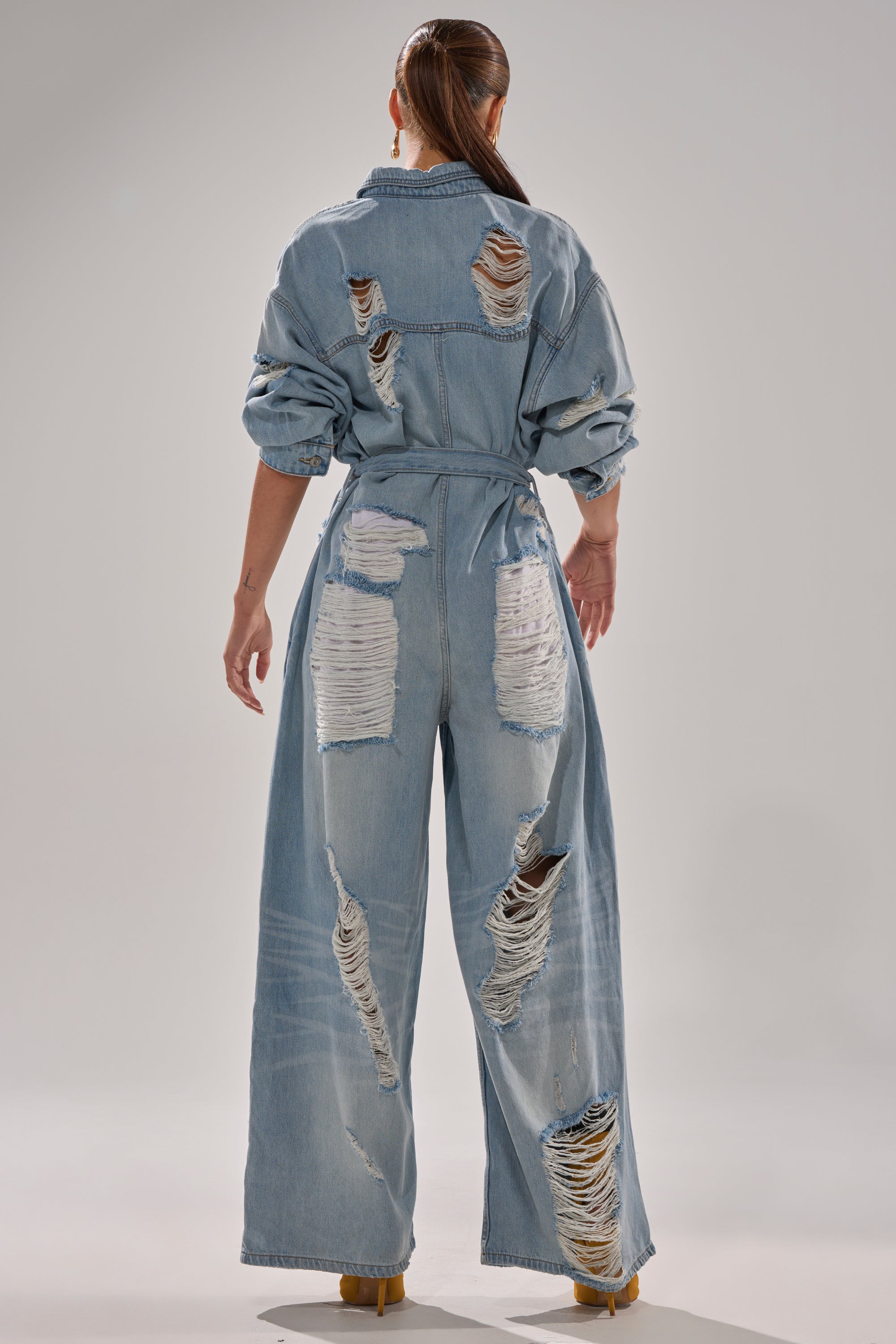 A woman with long brown hair in a ponytail models the NOT TODAY EXTREME OVERSIZED DISTRESSED DENIM JUMPSUIT, featuring large frayed rips on the back, arms, and wide legs. She stands facing away in a well-lit studio.