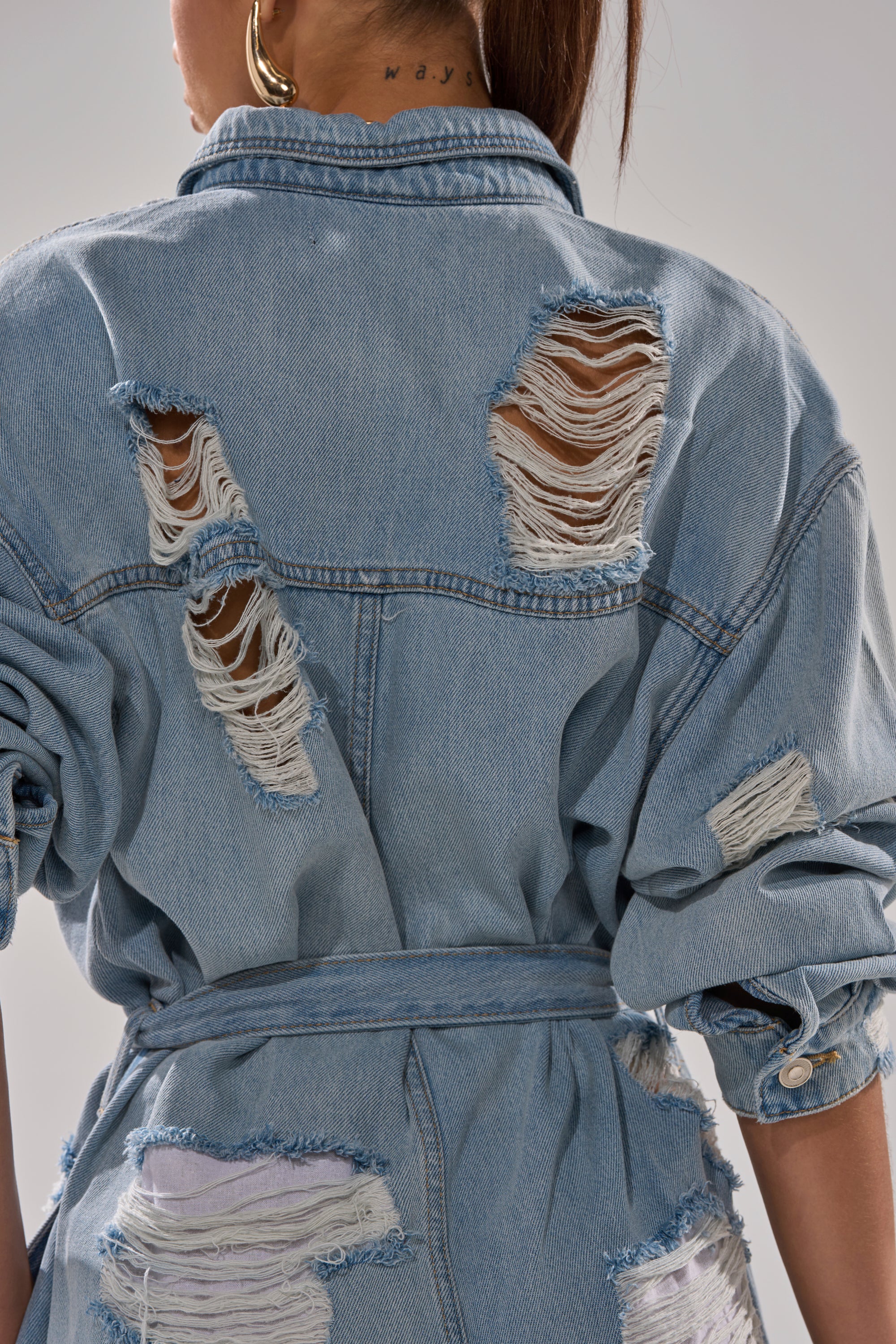 Wearing the NOT TODAY EXTREME OVERSIZED DISTRESSED DENIM JUMPSUIT in light blue, featuring large frayed rips on the back and sleeves, this belted piece creates an effortlessly edgy look when styled with long hair and a visible neck tattoo.