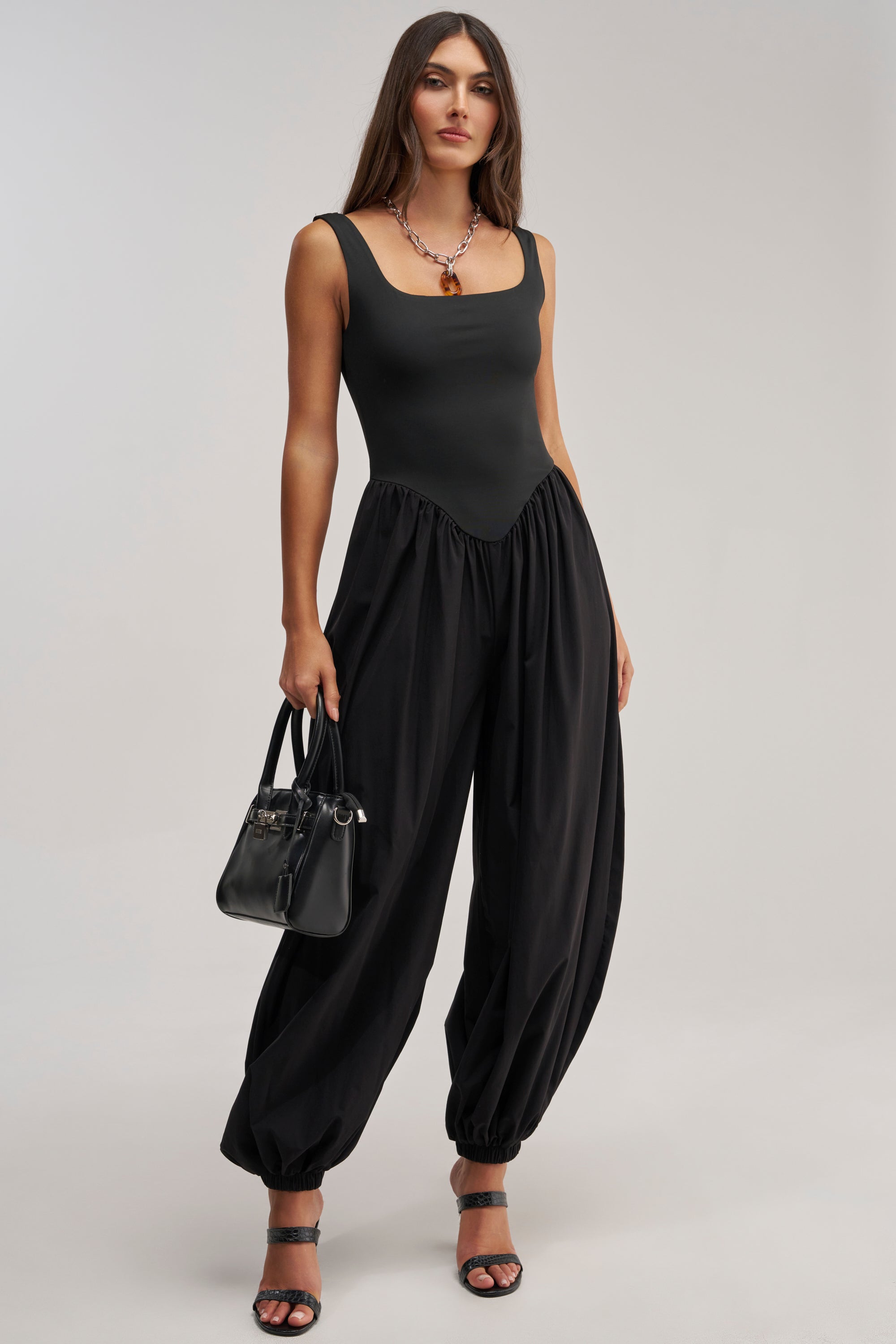 A woman with long brown hair wears the FLIRT ALERT SQUARE NECK JUMPSUIT and black heeled sandals, holding a black handbag. She stands against a plain light background, creating an effortlessly chic look.
