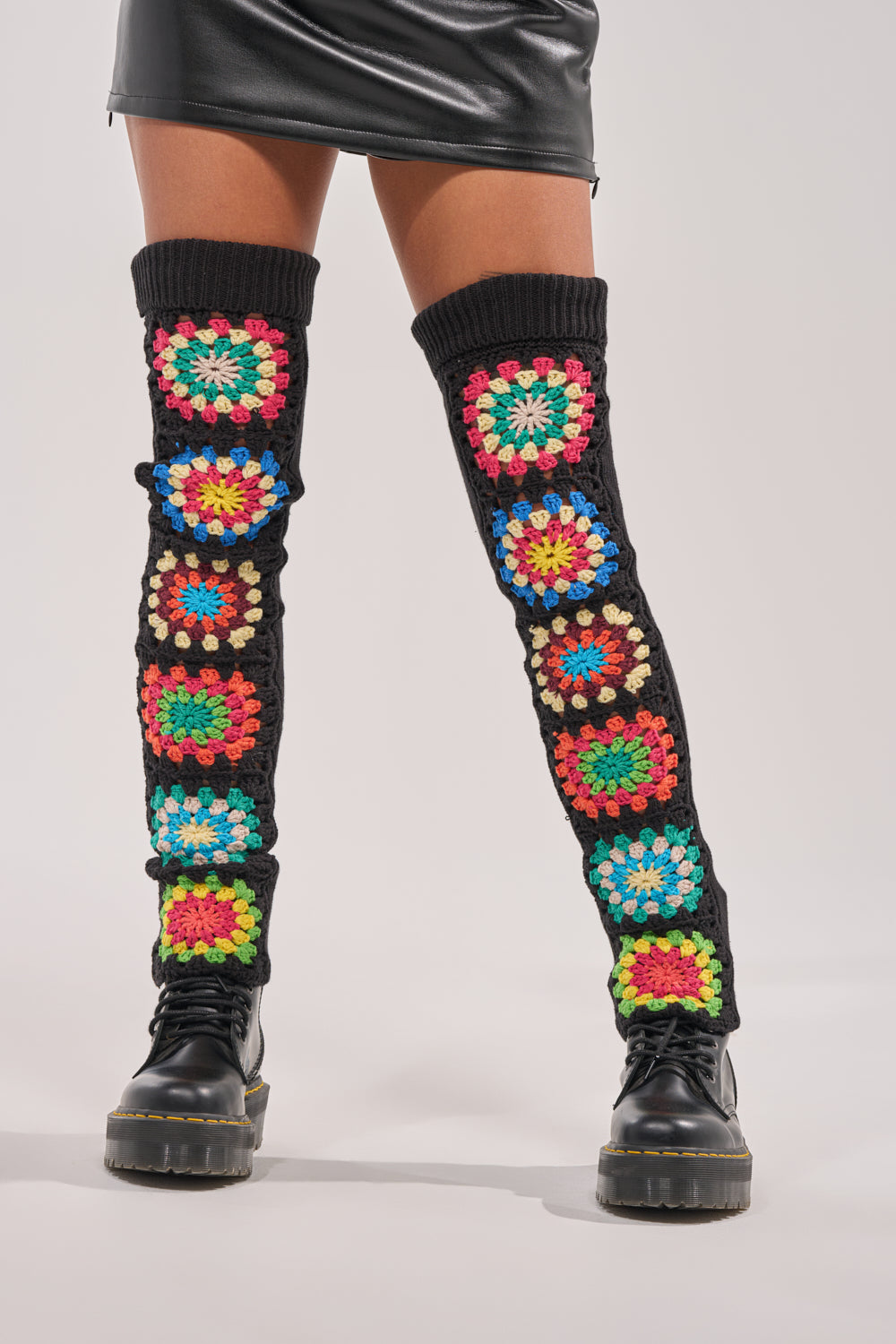 Someone wears TO THE BEACH SOCKS—rainbow crochet leg warmers with multicolored granny squares—paired with a black leather mini skirt and chunky black boots, standing against a plain, light-colored background. 0