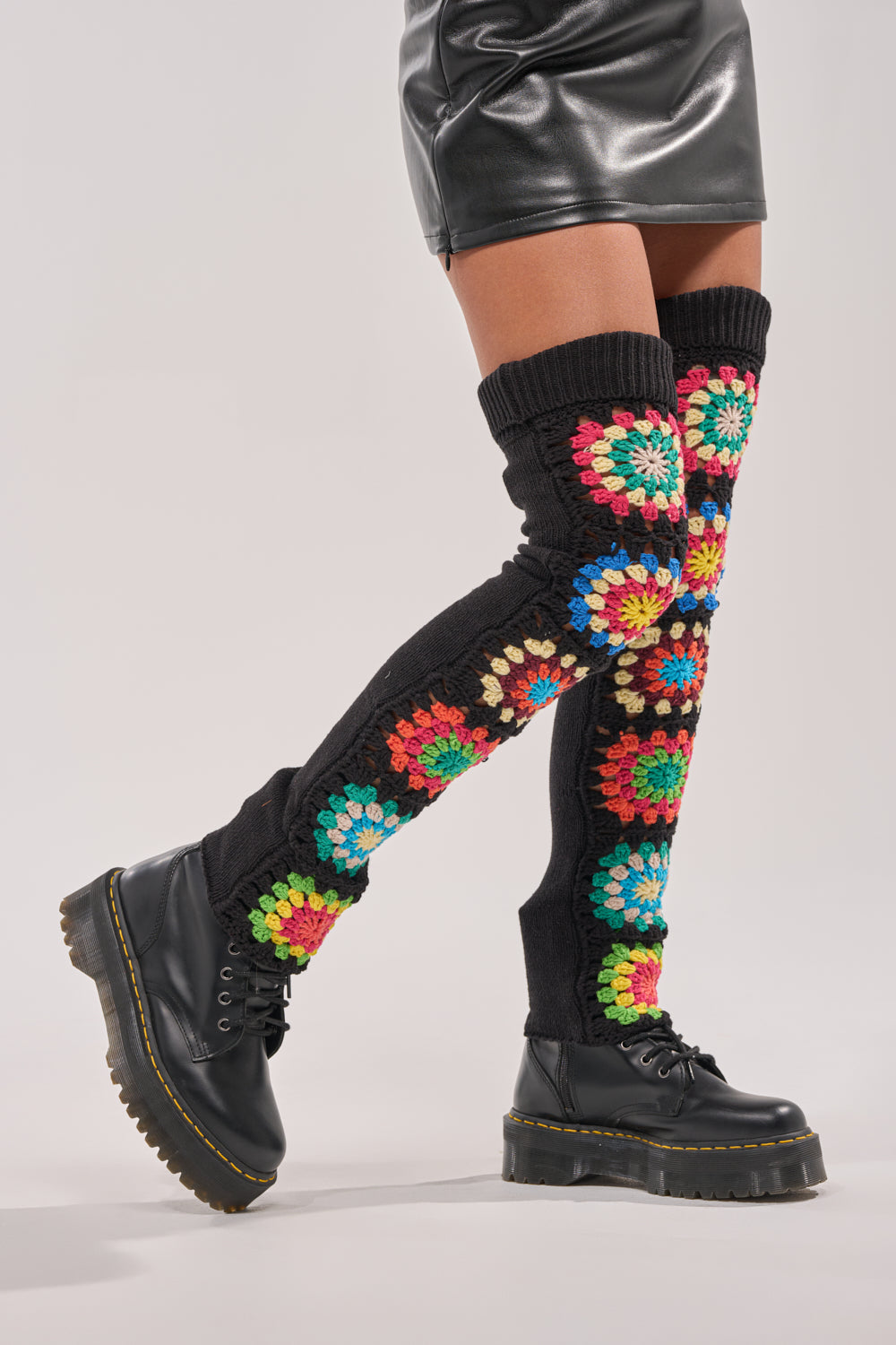 Someone wearing TO THE BEACH SOCKS—retro-inspired legwarmers with colorful floral crochet patterns—along with a short black leather skirt and black chunky lace-up boots on a plain light background. 1