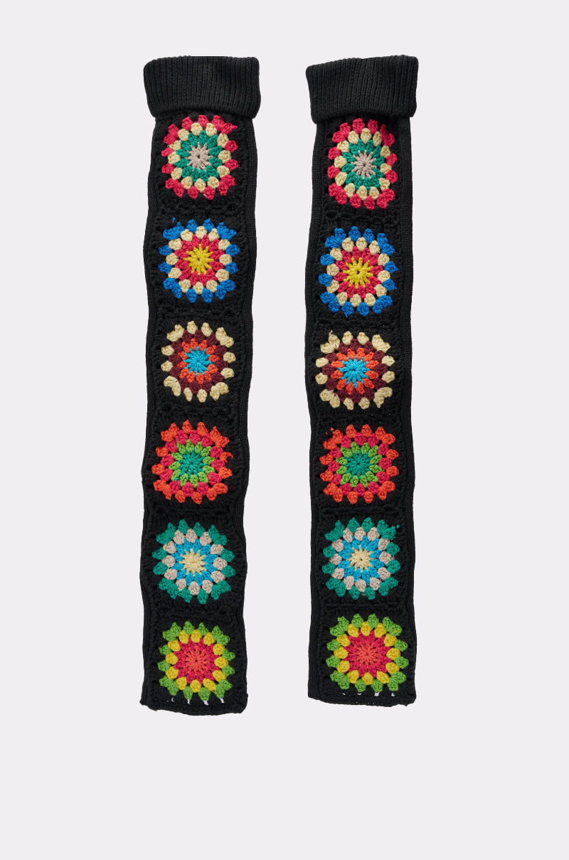 TO THE BEACH SOCKS: A pair of black crocheted leg warmers, each adorned with a row of rainbow crochet floral patterns in red, blue, yellow, green, and beige. Set against a plain white background. 2