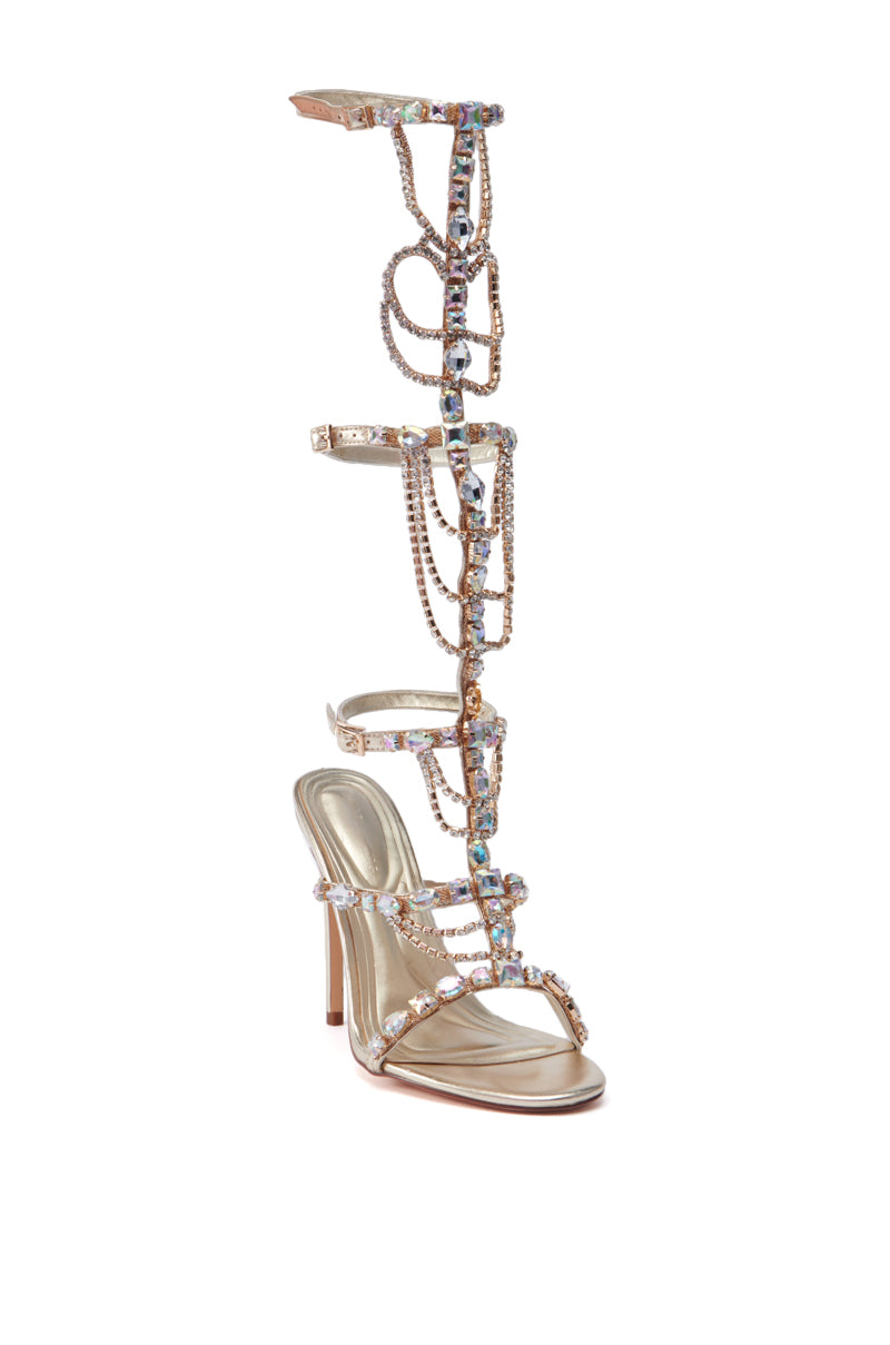 AZALEA WANG REALEM gold embellished gladiator sandal featuring multiple straps adorned with sparkling gemstones, extending up the leg with additional decorative chains and a sleek stiletto heel.