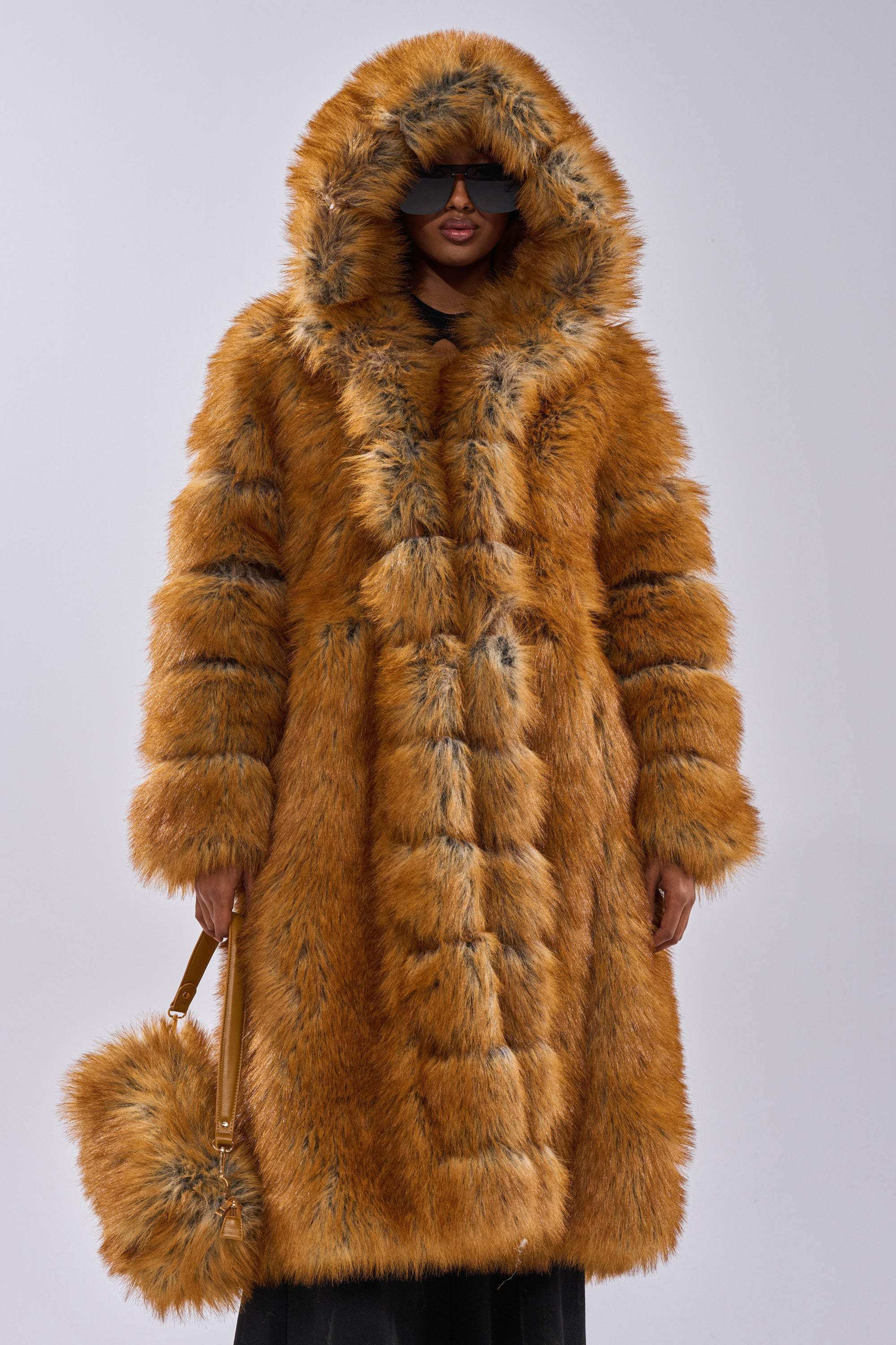 A person embodies old money elegance in the OLD MONEY FAUX FUR COAT—a large, hooded faux fur coat—paired with a matching furry purse and dark sunglasses, posed against a plain light background.