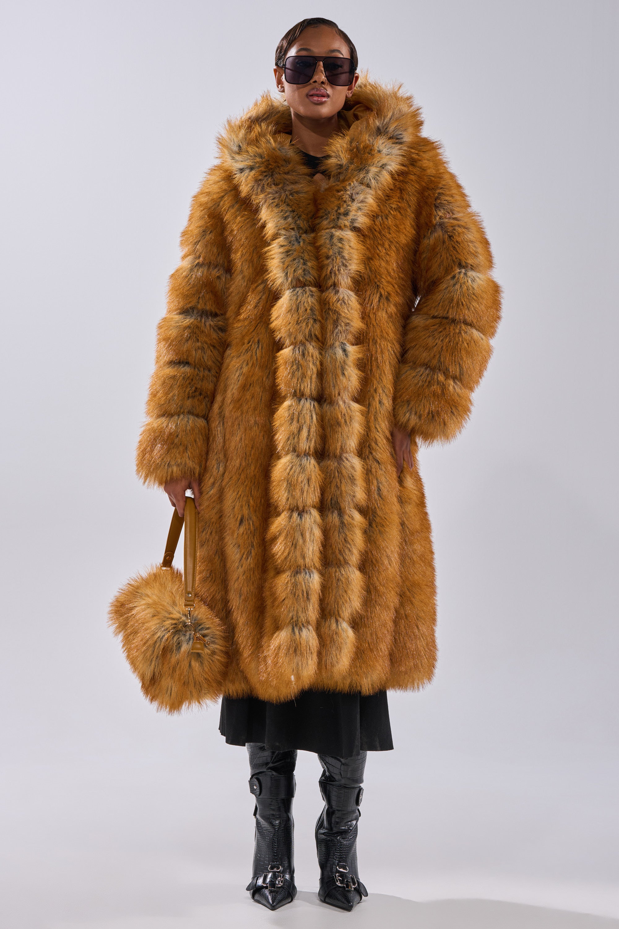 Wearing the OLD MONEY FAUX FUR COAT in orange with a hood, oversized sunglasses, black boots, and a matching fur handbag, this person radiates old money style against a plain light background.