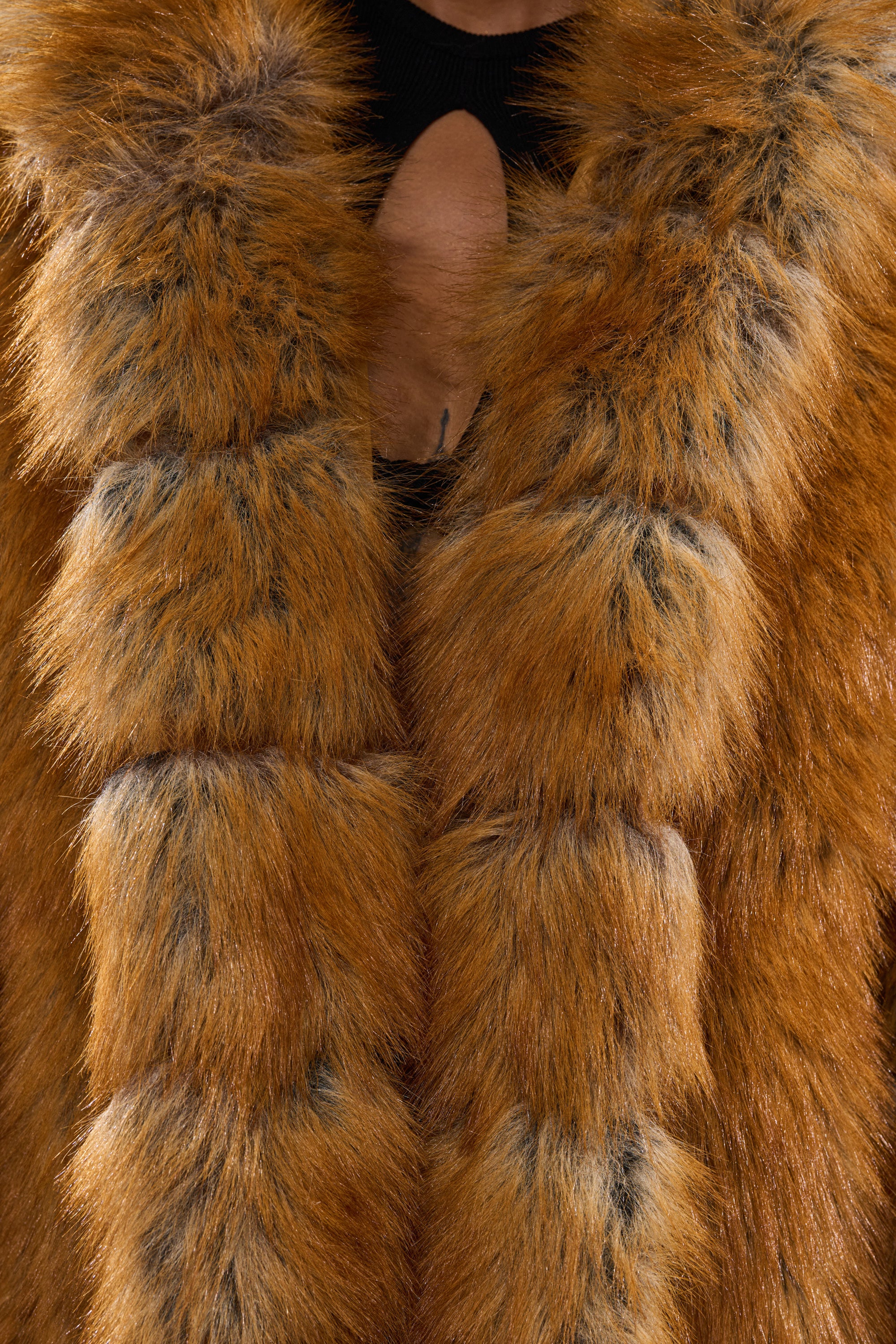 Close-up of a person in the OLD MONEY FAUX FUR COAT, a thick orange-brown faux fur piece with black accents, worn over a black top with a round chest cutout. Only the lower face and neck are partially visible.