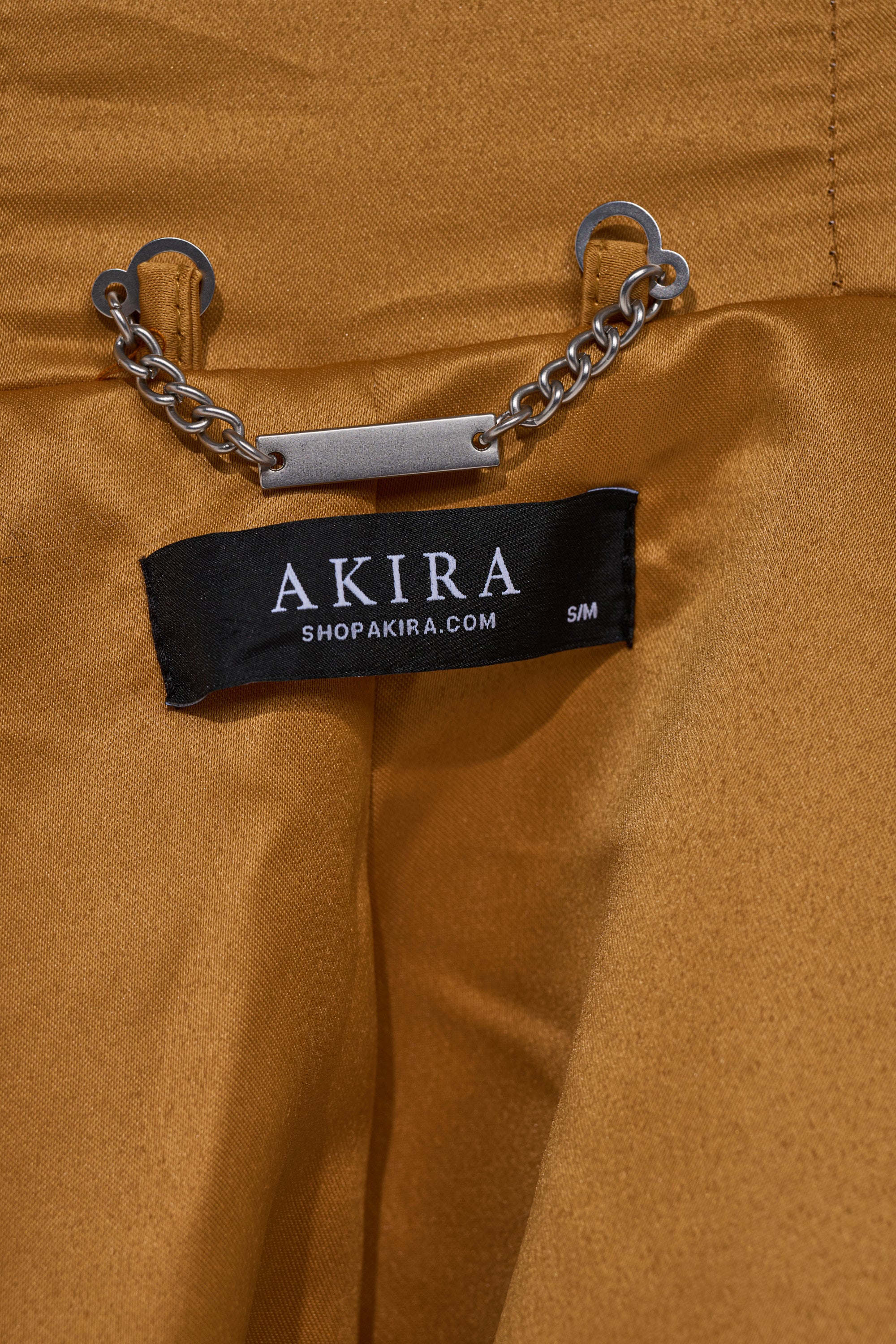 A close-up of the mustard-yellow OLD MONEY FAUX FUR COAT displays a silver chain with a metal bar and a black "AKIRA SHOPAKIRA.COM S/M" label.