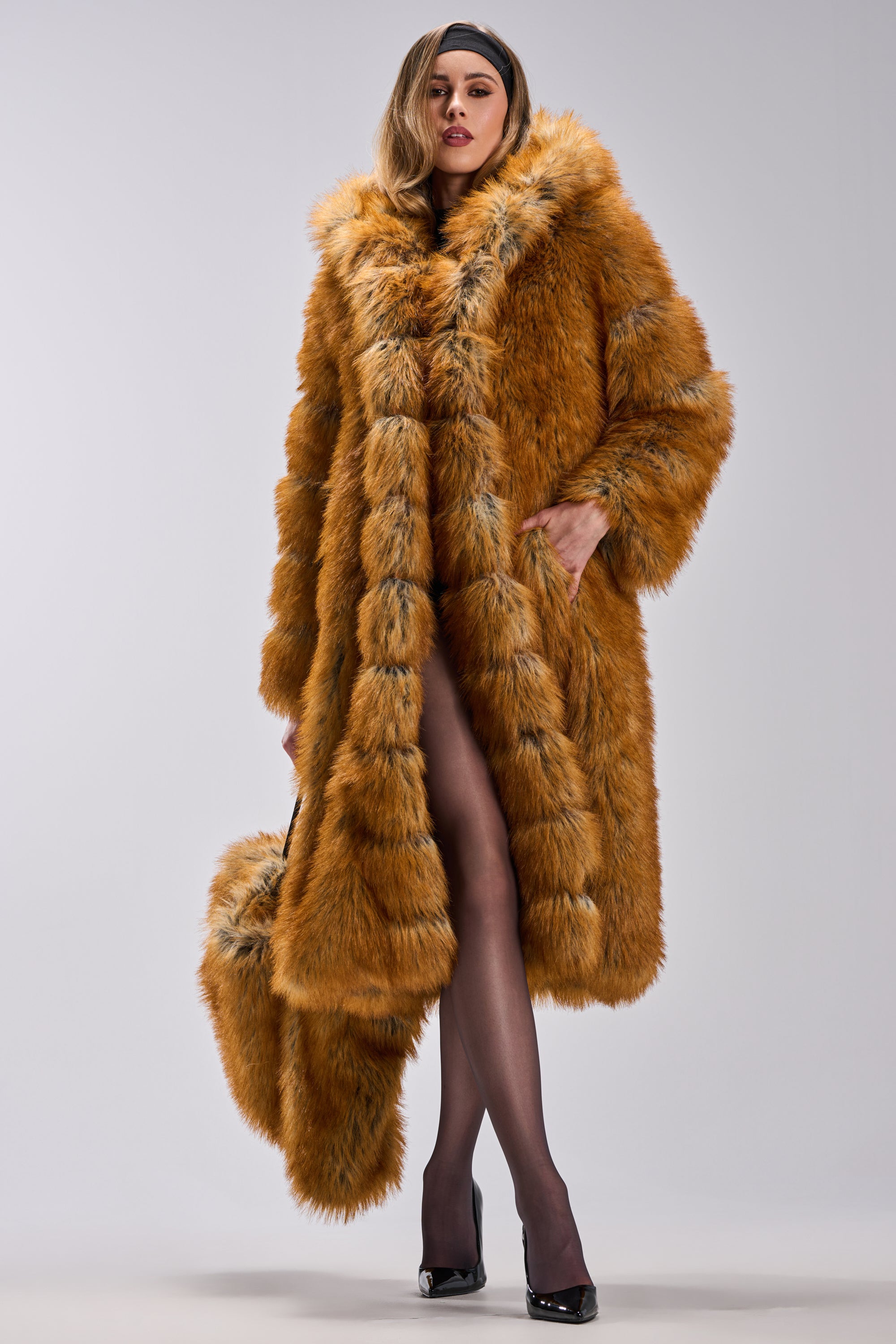 A woman stands confidently in the OLD MONEY FAUX FUR COAT with a hood, holding a matching fur bag. She pairs it with black sheer stockings and high heels, set against a plain light gray background.