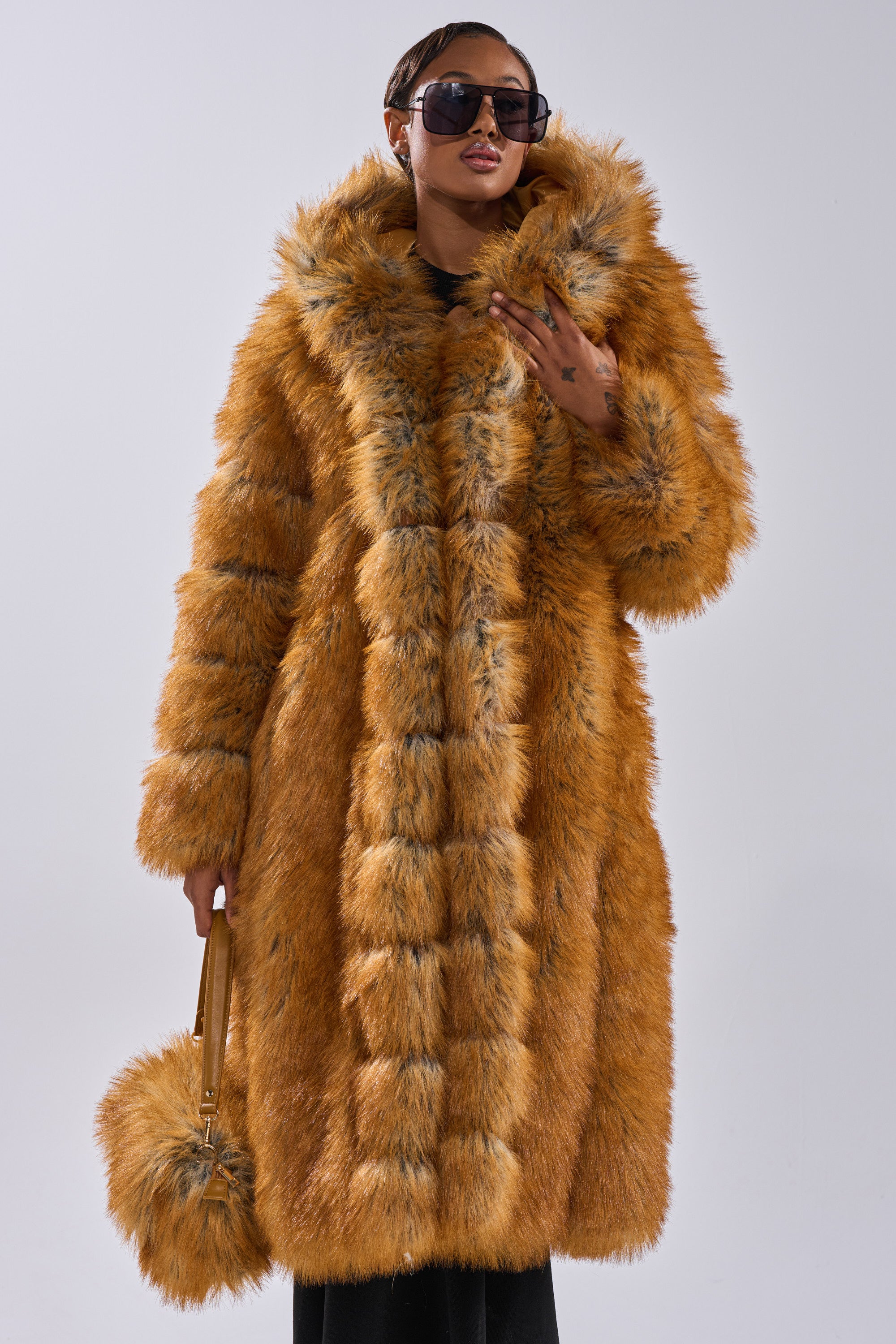 A person in the OLD MONEY FAUX FUR COAT, with large sunglasses, stands against a plain background, holding a matching round fur handbag and touching their sunglasses.