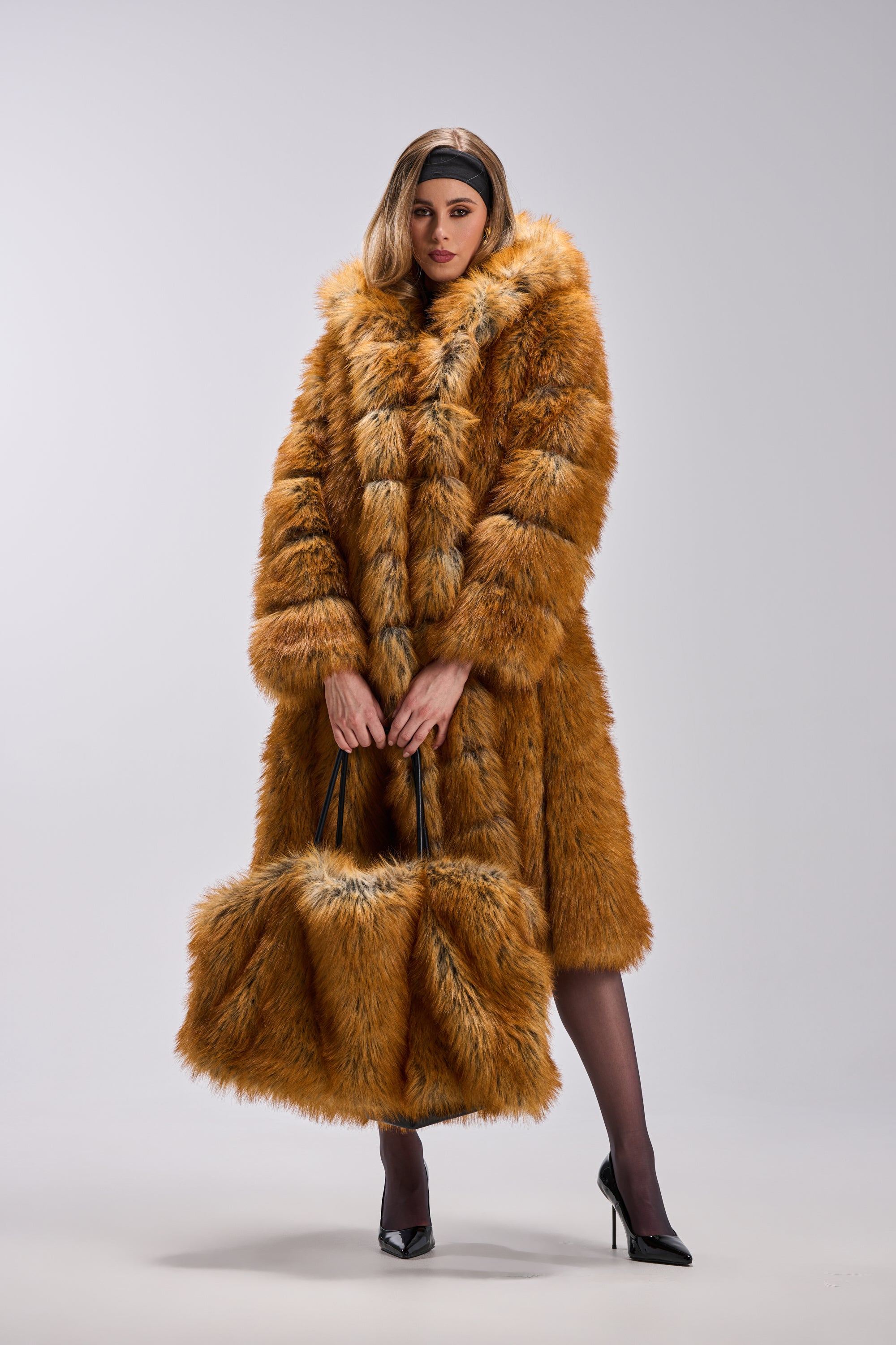 Dressed in the OLD MONEY FAUX FUR COAT—a long, fluffy brown piece—she carries a matching large fur bag, styled with a black headband, sheer tights, and high heels against a light backdrop.