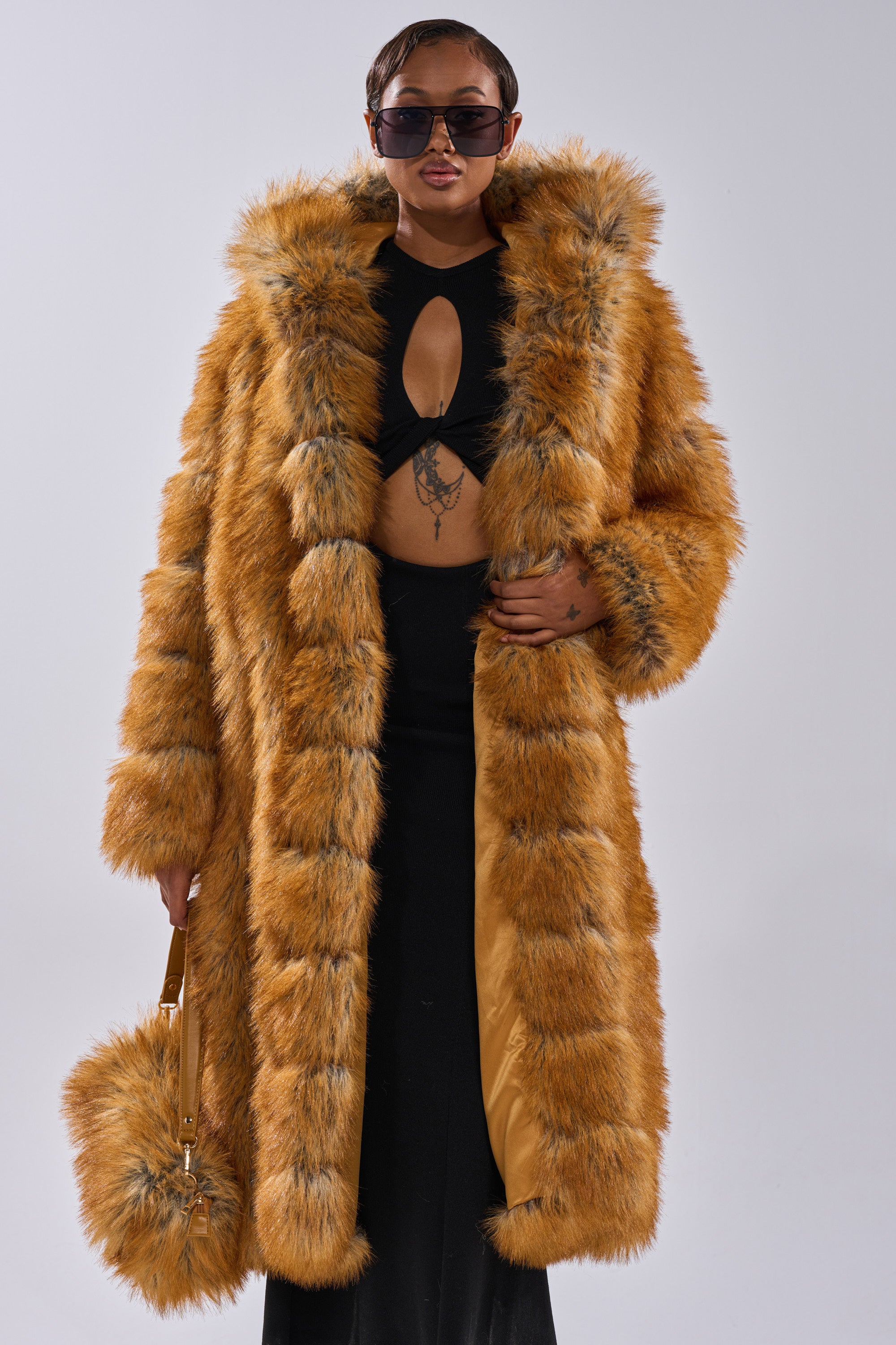 Wearing the OLD MONEY FAUX FUR COAT—an elegant orange piece with black accents—a person pairs it with dark sunglasses, a matching furry handbag, and a black cutout dress, standing against a plain light background.