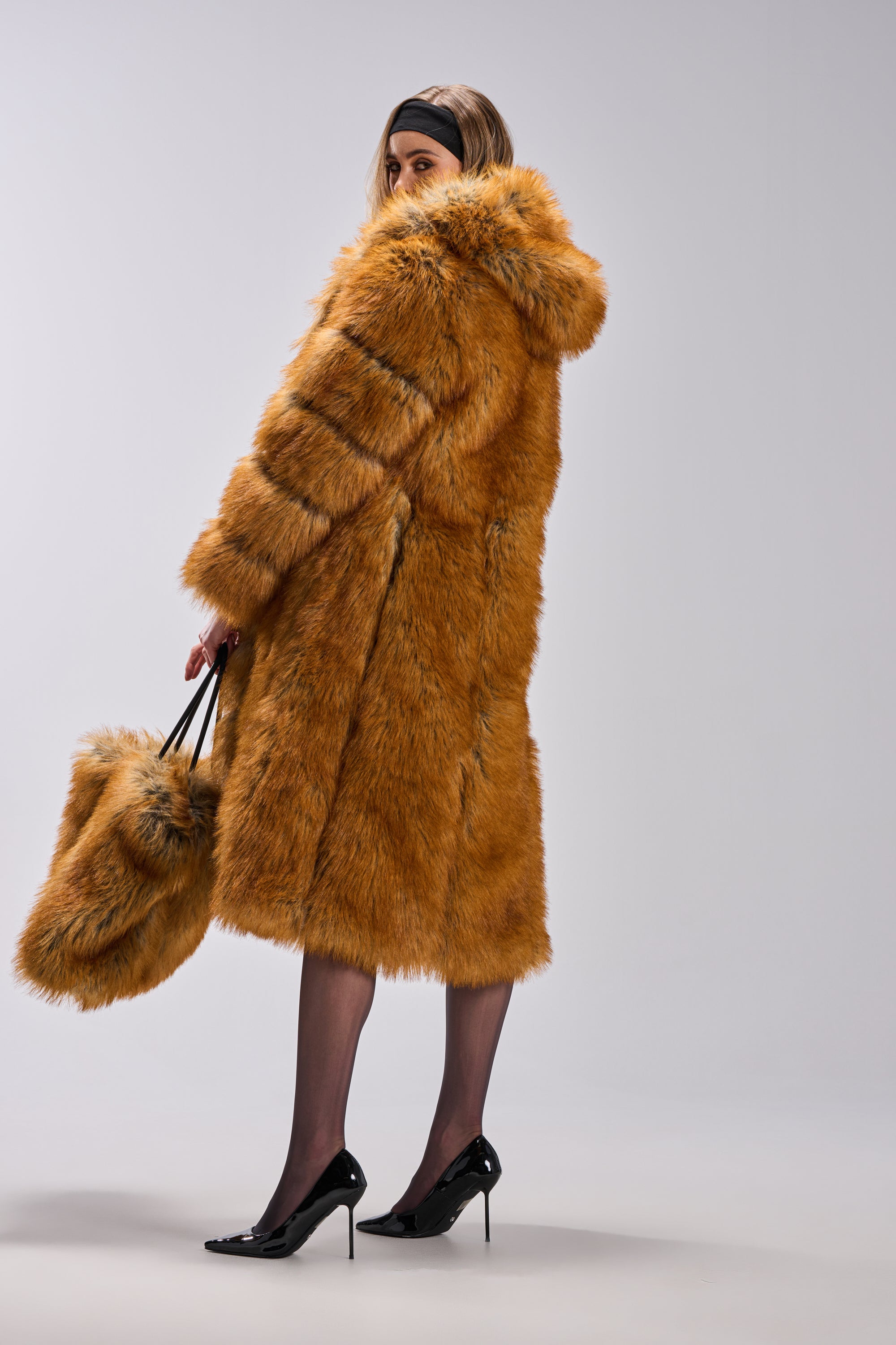 A person models the OLD MONEY FAUX FUR COAT—long, orange, with a hood—and holds a matching bag while standing in black high heels against a plain light gray background, highlighting this luxury outerwear piece.