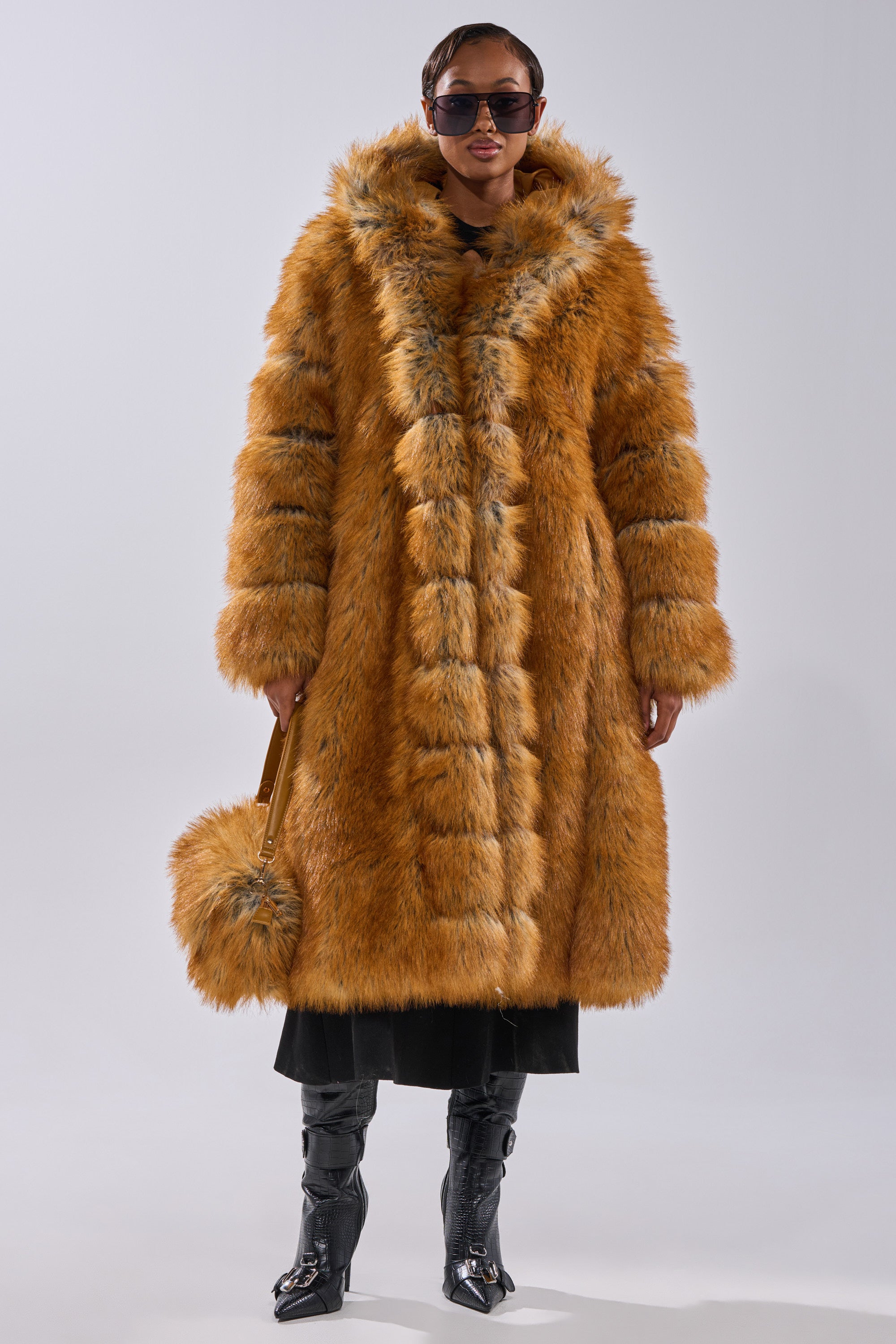 Wearing sunglasses, shiny black boots, and a matching furry bag, a person in the OLD MONEY FAUX FUR COAT—a long, fluffy orange hooded coat—pairs it with a black skirt or pants and stands against a plain light background.