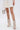 A person wears a white skirt and AZALEA WANG REEVES NEW OPEN TOE PU BOOTIE IN BONE with stiletto heels, holding a small silver geometric handbag against a plain white background.