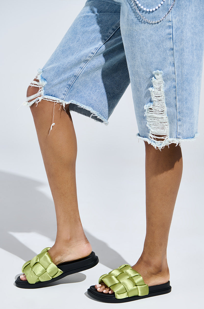 A person in light blue, knee-length ripped denim shorts with frayed edges stands against a plain background, wearing AKIRA REIGNS Olive Braided Puff Slip-On Sandals featuring thick, padded straps for effortless style.