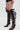 A person wears black faux leather shorts and the AZALEA WANG REIN BLACK THIGH HIGH BOOT, featuring wide cut-out straps and stiletto heels, for a bold modern look against a plain light-colored background.