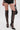 A person stands against a plain white background, wearing black faux leather shorts and the AZALEA WANG REIN BLACK THIGH HIGH BOOT with stiletto heels, and holding a small black handbag.