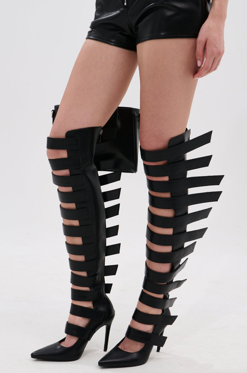 A person wears the AZALEA WANG REIN BLACK THIGH HIGH BOOT, featuring striking stiletto heels, multiple horizontal cutouts, and wide straps. They are styled with black faux leather shorts against a plain white background.