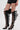 A person wears the AZALEA WANG REIN BLACK THIGH HIGH BOOT, featuring striking stiletto heels, multiple horizontal cutouts, and wide straps. They are styled with black faux leather shorts against a plain white background.