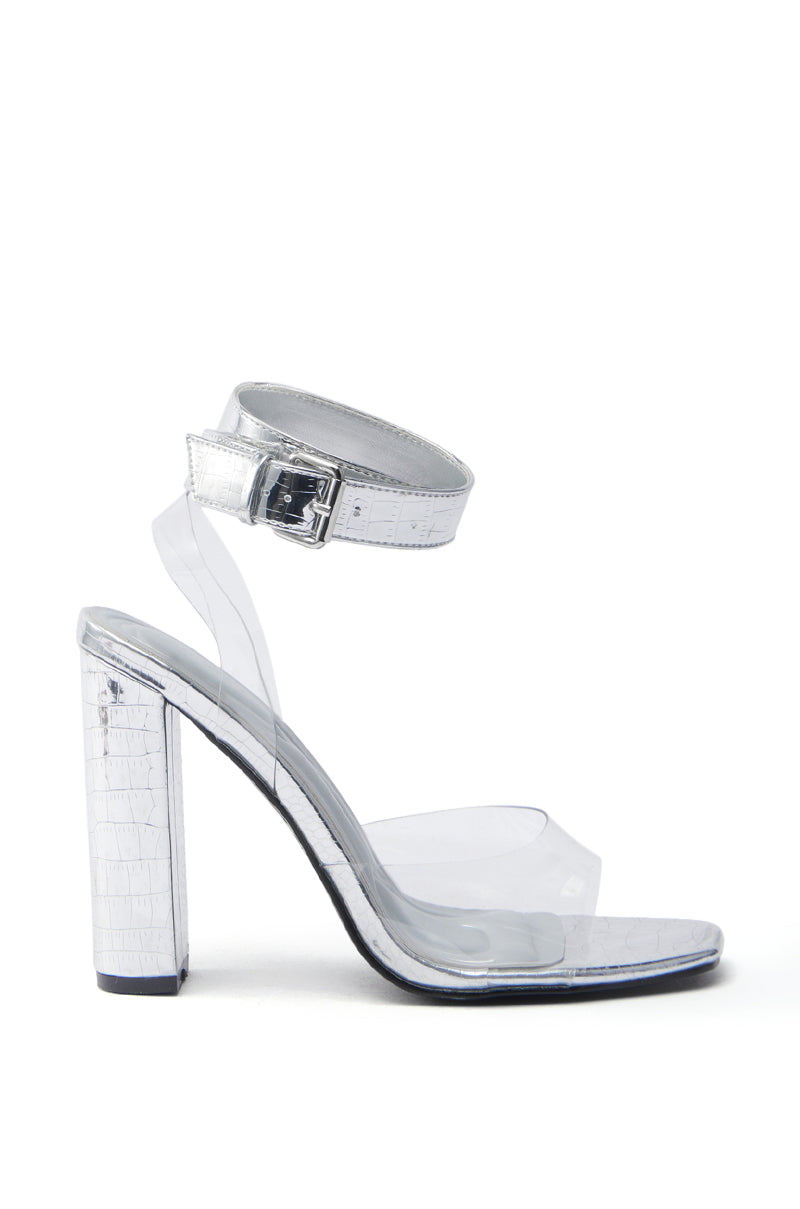 The AZALEA WANG REMILIA SILVER CROC SANDAL WITH STRAP features a clear high heel and transparent ankle strap with a silver buckle, showcasing a thick textured heel and sole for a sleek, modern look against a white background.