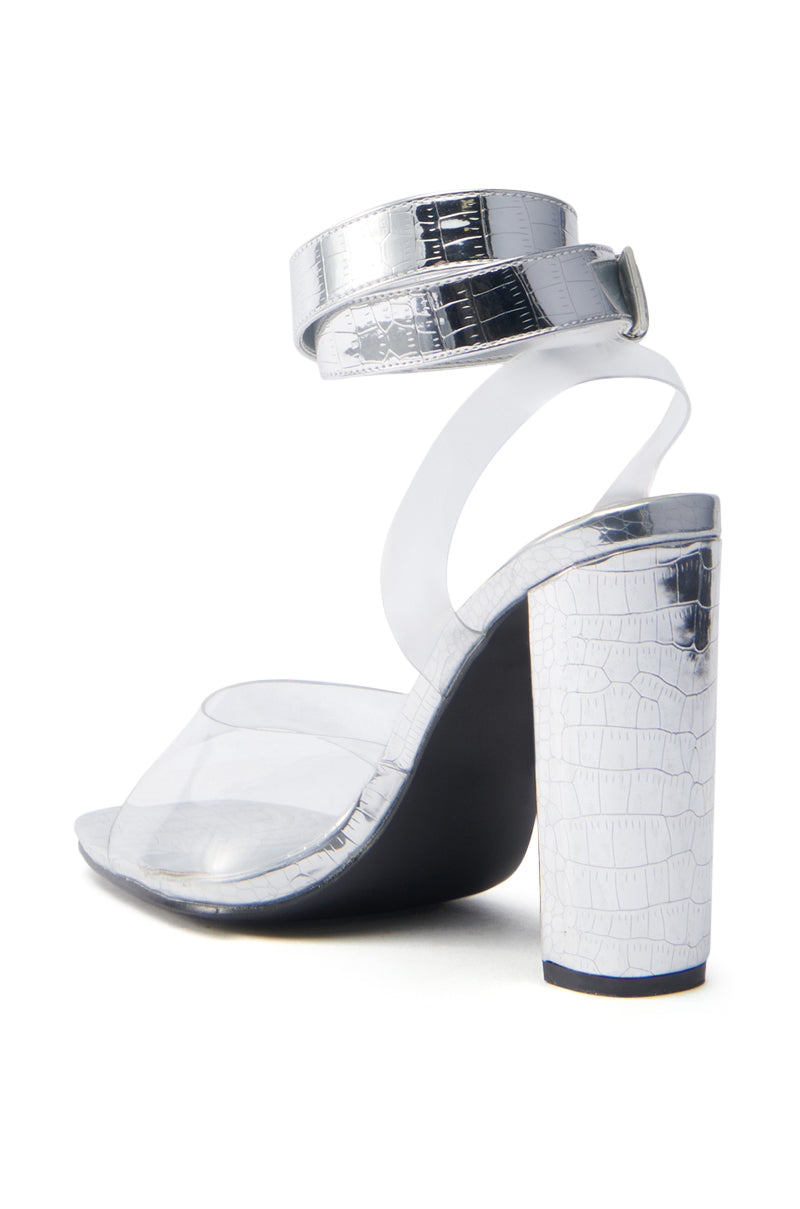 The AZALEA WANG REMILIA SILVER CROC SANDAL features a chunky silver croc-textured heel, an ankle strap, a glossy faux leather finish, and a clear strap across the toes. The image provides a back and side view of this strappy high-heel shoe.