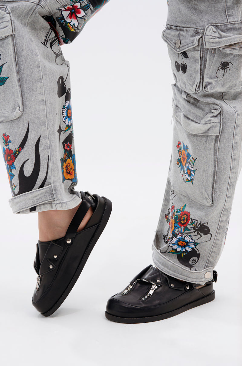 Close-up of a person in light gray jeans with colorful prints, paired with AZALEA WANG RESTER BLACK HARDWARE EMBELLISHED MULE shoes featuring metallic zipper details, against a plain white background.
