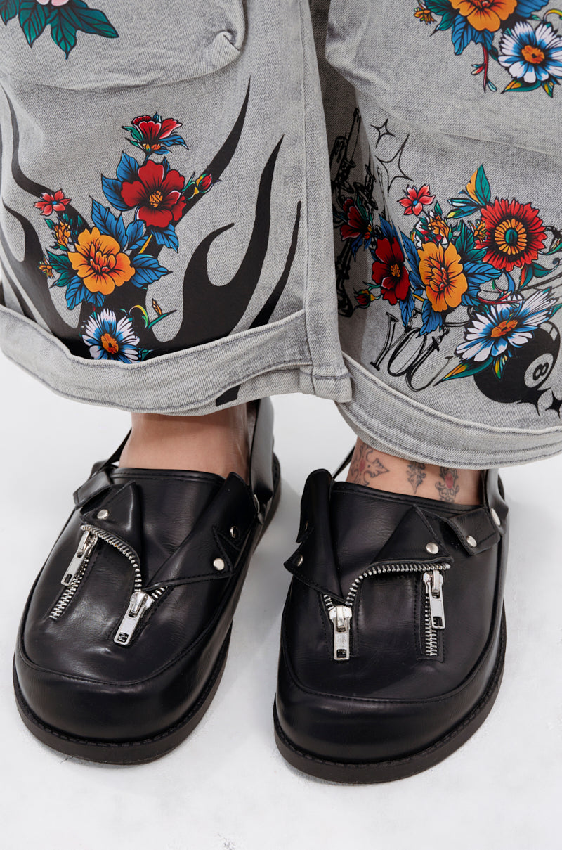 Close-up of feet in AZALEA WANG RESTER BLACK HARDWARE EMBELLISHED MULES with metallic zipper detail, worn with gray floral-patterned pants and visible tattooed ankles above the shoes.