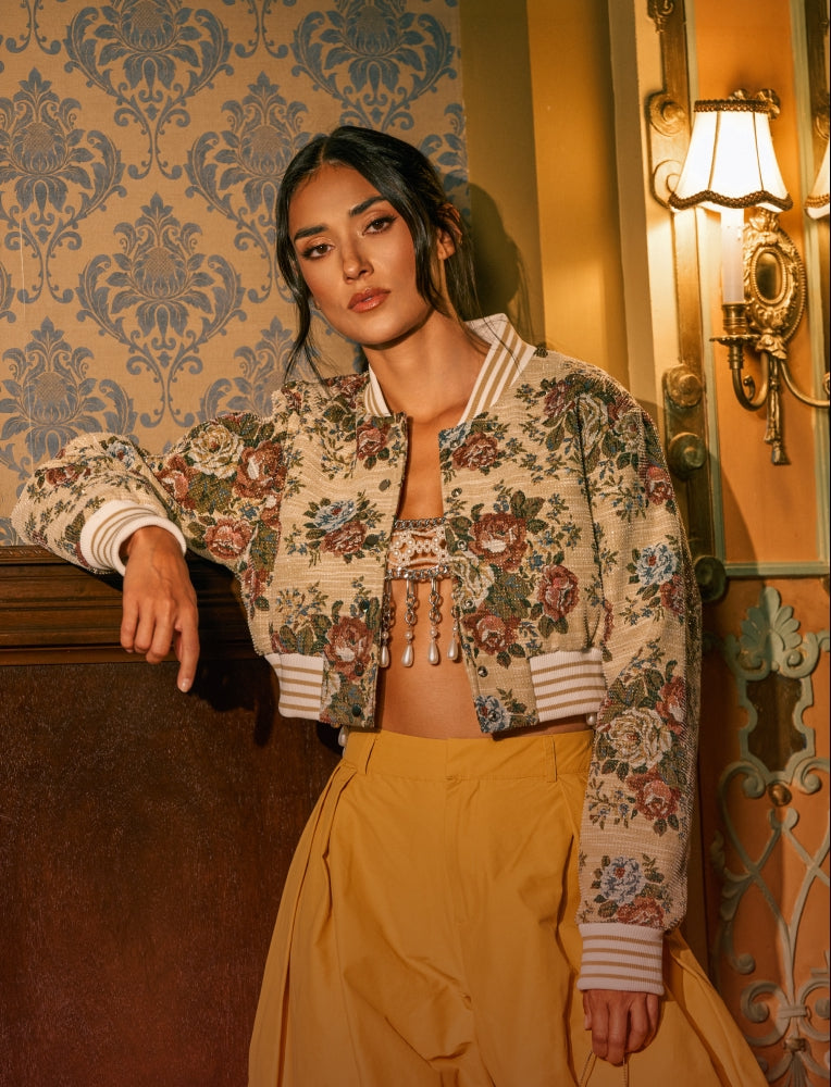 A woman models a cropped floral bomber jacket with a bohemian tassel detail at the center, paired with a mustard yellow skirt, set against a vintage-style wallpaper and wall sconce background.