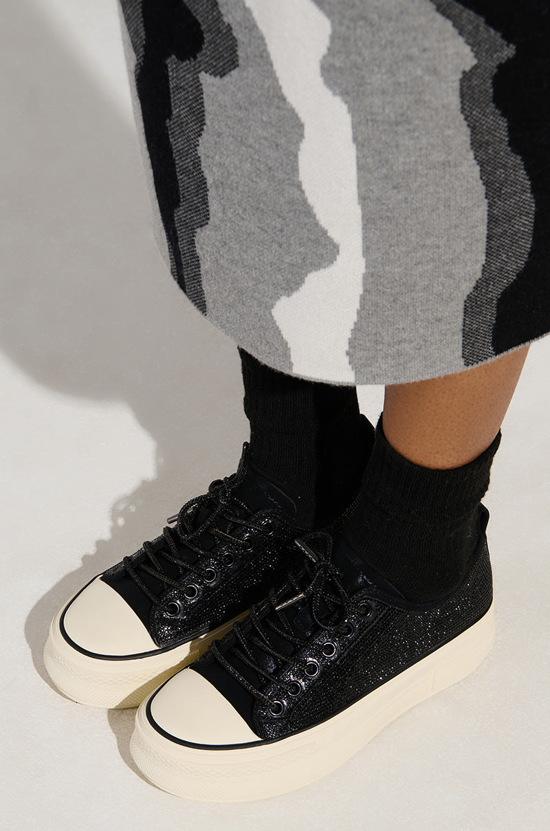 A person wearing the AZALEA WANG REVIVAL BLACK RHINESTONE FLATFORM SNEAKER with white soles and a faux leather upper, complemented by black socks and a skirt with a black, white, and gray abstract pattern, stands on a light surface.