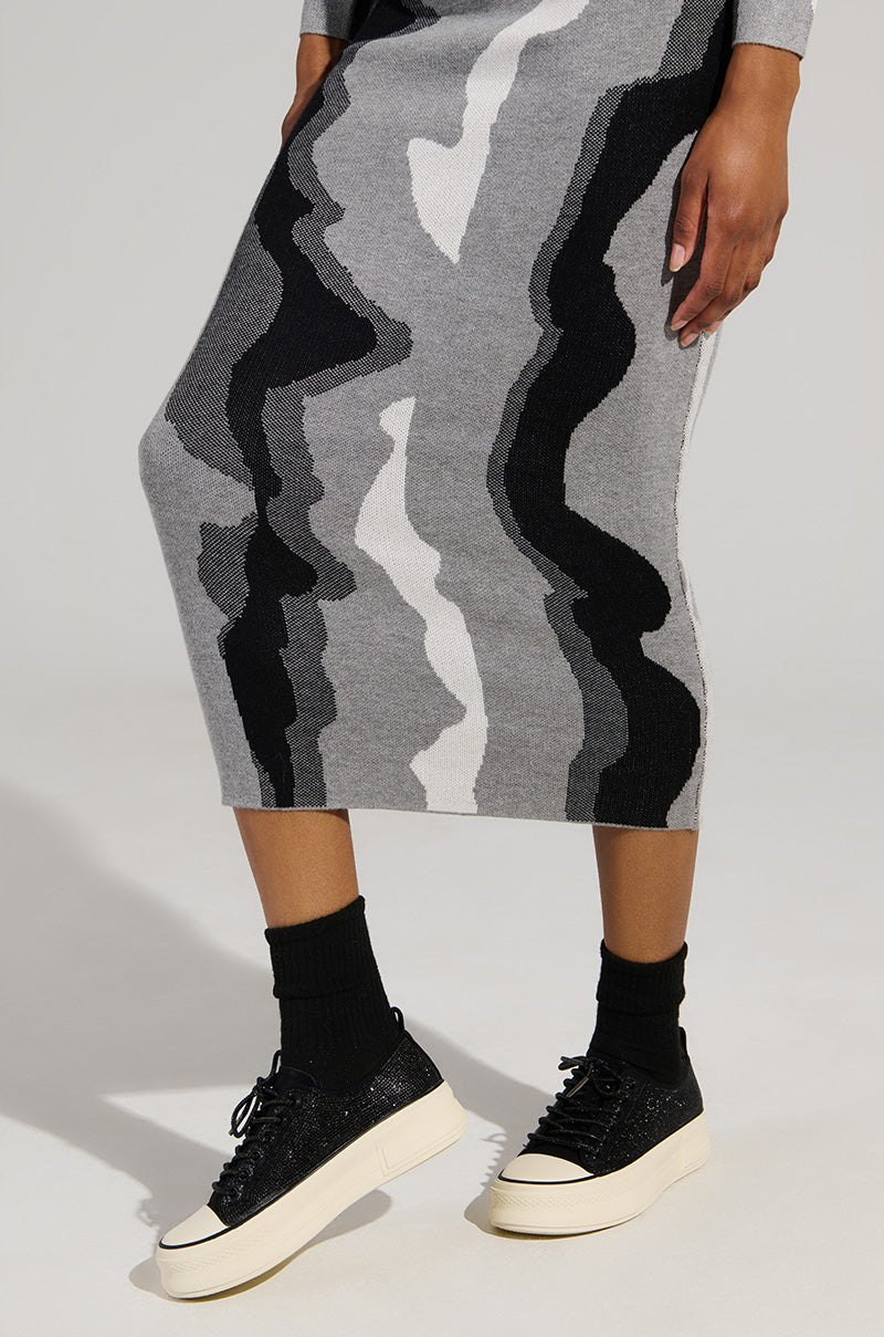 The individual is dressed in a long, wavy-patterned skirt in shades of black, white, and gray, complemented by black socks and the stylish AZALEA WANG REVIVAL BLACK RHINESTONE FLATFORM SNEAKERS. These sneakers are adorned with faux leather uppers and are set on eye-catching white flatform soles that stand out against a simple light backdrop.