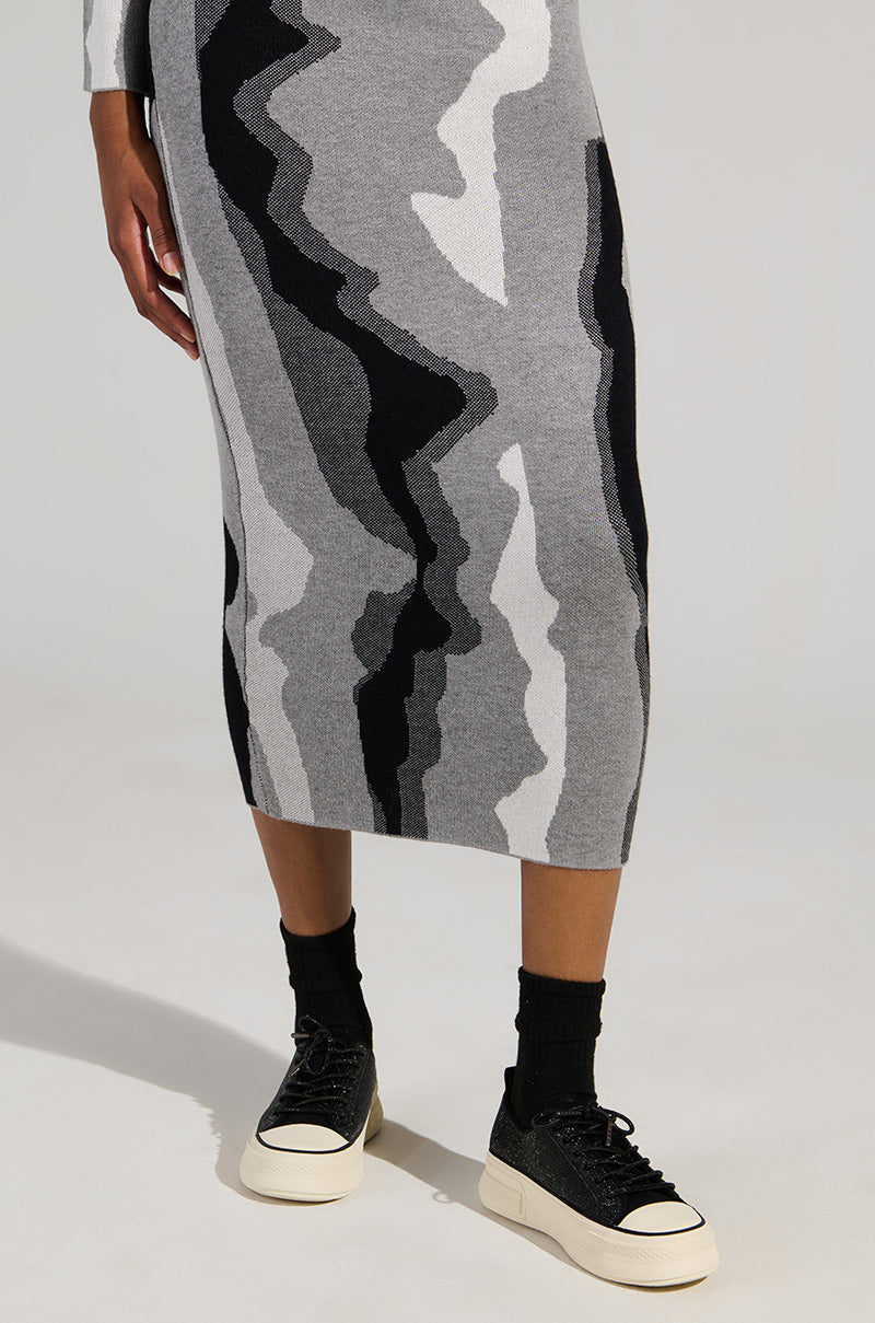 A person wearing a gray dress featuring abstract black and white patterns is standing, displaying only their lower body. They are sporting the chic AZALEA WANG REVIVAL BLACK RHINESTONE FLATFORM SNEAKERS along with black socks against a plain background.