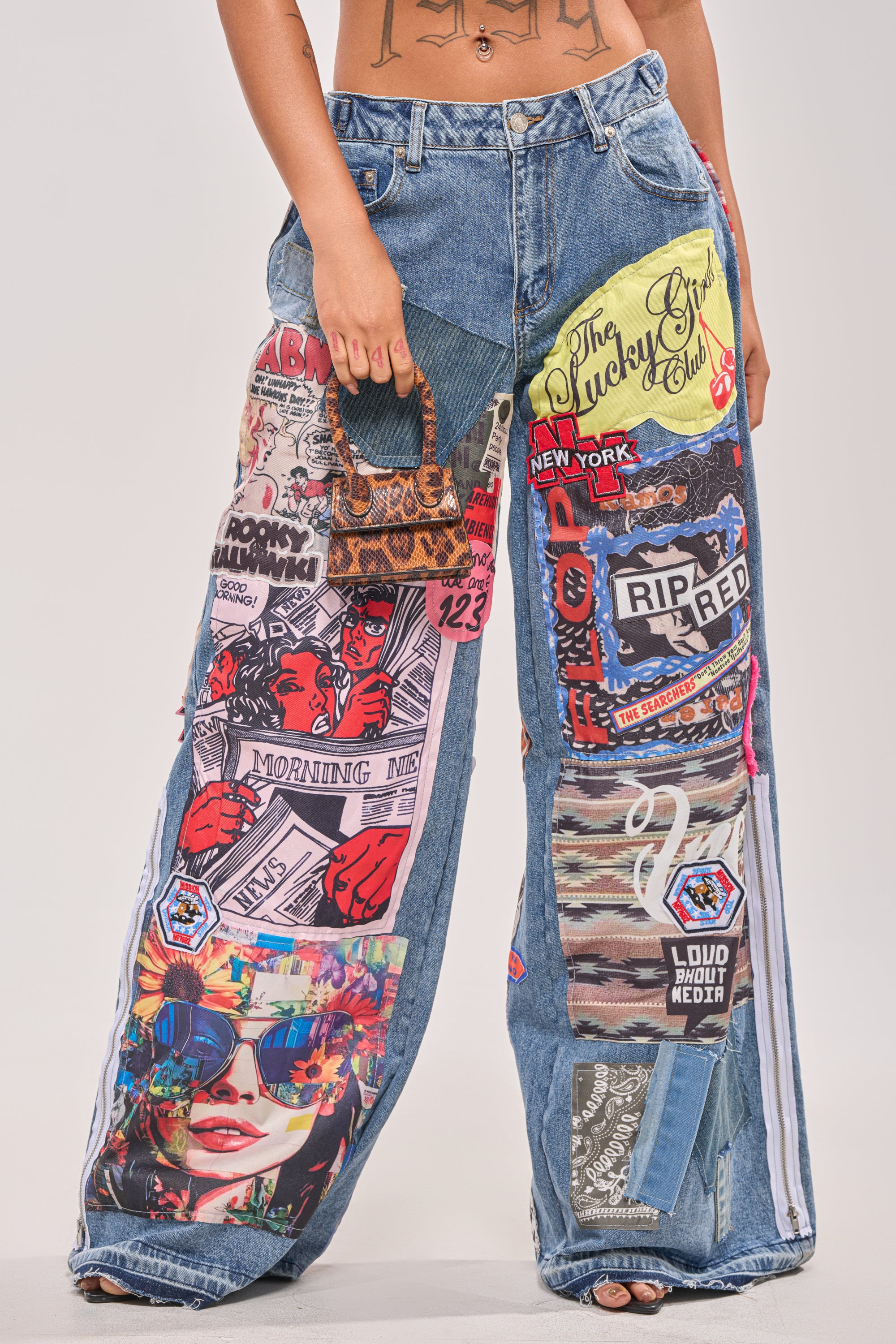 A person wears the SO SICK SO COOL VERY OVERSIZED DENIM, featuring colorful patches, comic-style graphics, and text. They carry a small patterned handbag. Only their torso and legs are shown against a plain background. 1