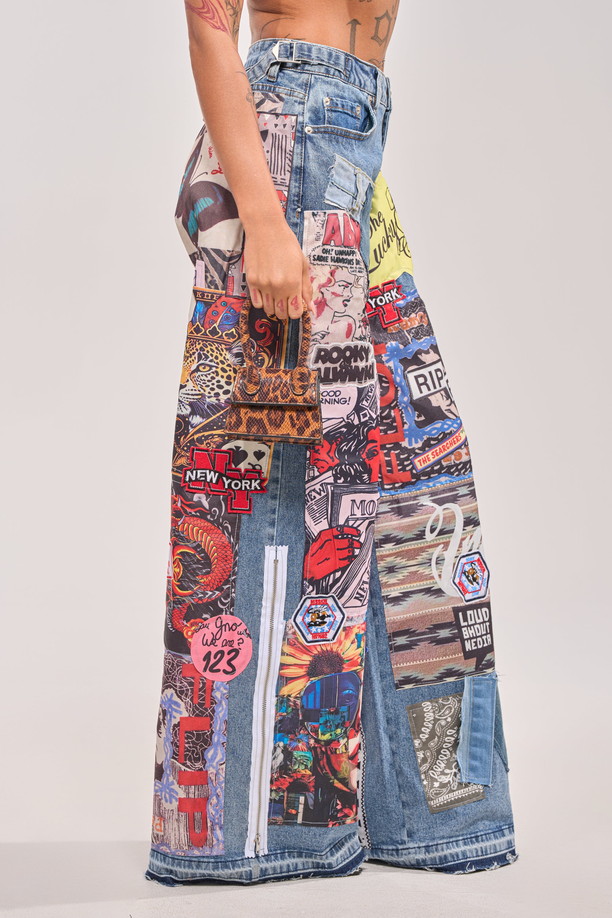 Person models SO SICK SO COOL VERY OVERSIZED DENIM, featuring ultra-oversized patchwork jeans with colorful eclectic patches, city names, images, and bold streetwear art. Only the lower torso and legs are shown. 2