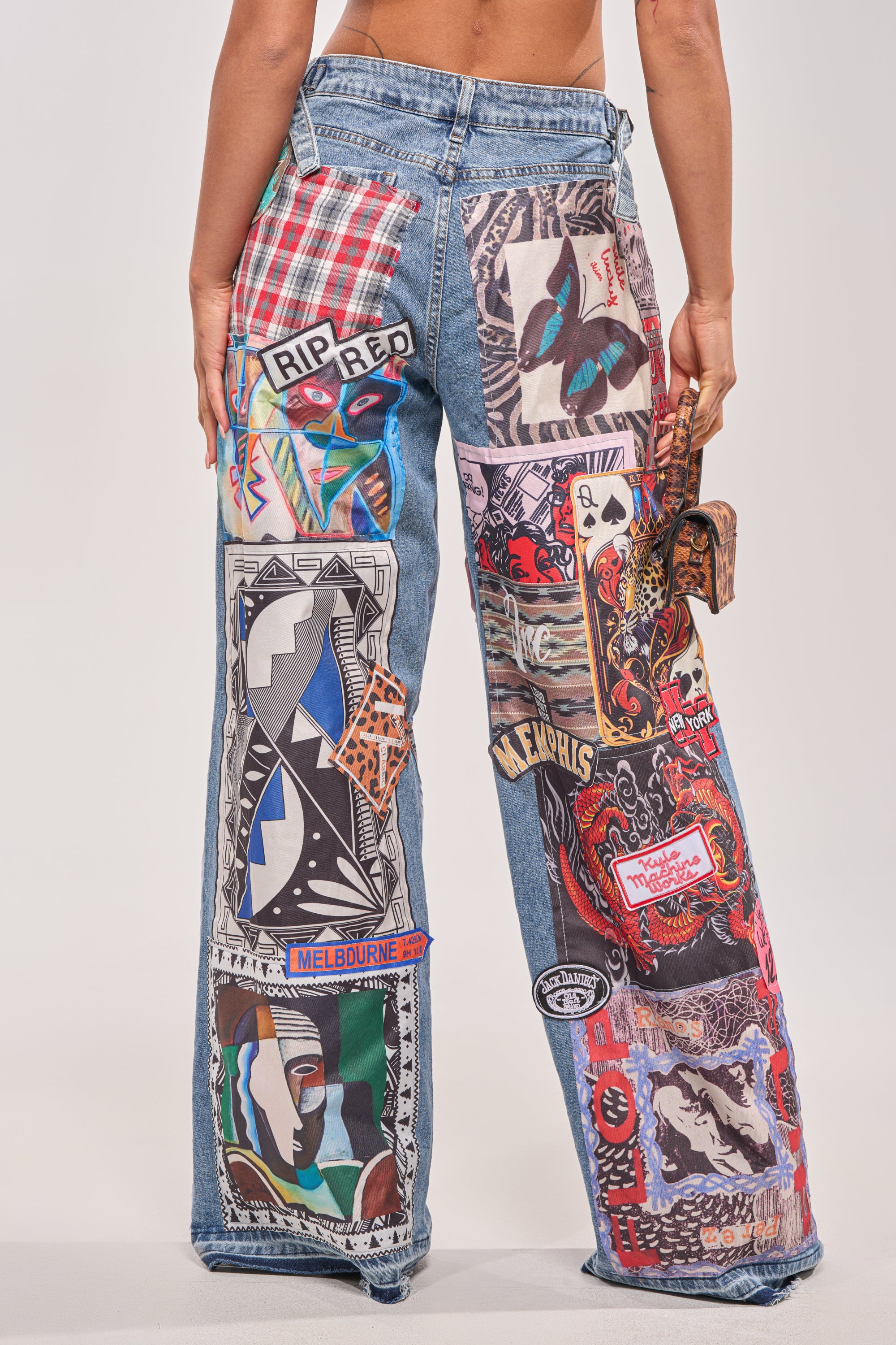 Someone wears the SO SICK SO COOL VERY OVERSIZED DENIM—ultra-wide leg jeans with colorful, patchwork fabric designs, text, and animal motifs. The photo is taken from behind to showcase the vibrant decorated streetwear legs. 4