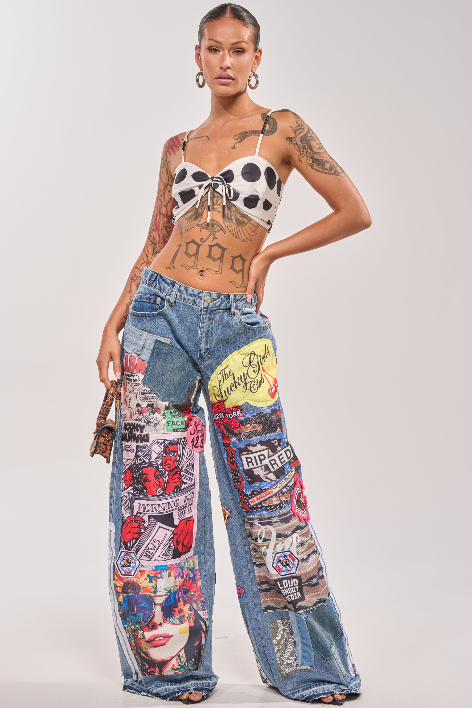A woman with tattoos and slicked-back hair rocks the SO SICK SO COOL VERY OVERSIZED DENIM jeans featuring colorful graphic patches, paired with a polka-dot crop top and gold hoop earrings for a bold streetwear vibe. 5
