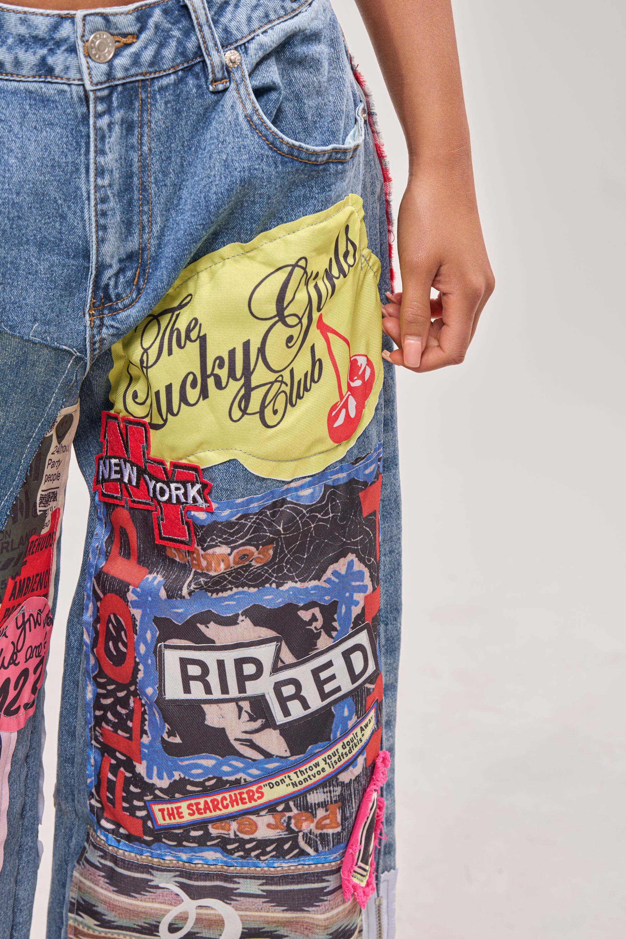 A person wears SO SICK SO COOL VERY OVERSIZED DENIM: ultra wide leg patchwork jeans with colorful designs and bold phrases like "The Lucky Guys Club," "NEW YORK," and "RIP RED." Their hand rests by their side. 7
