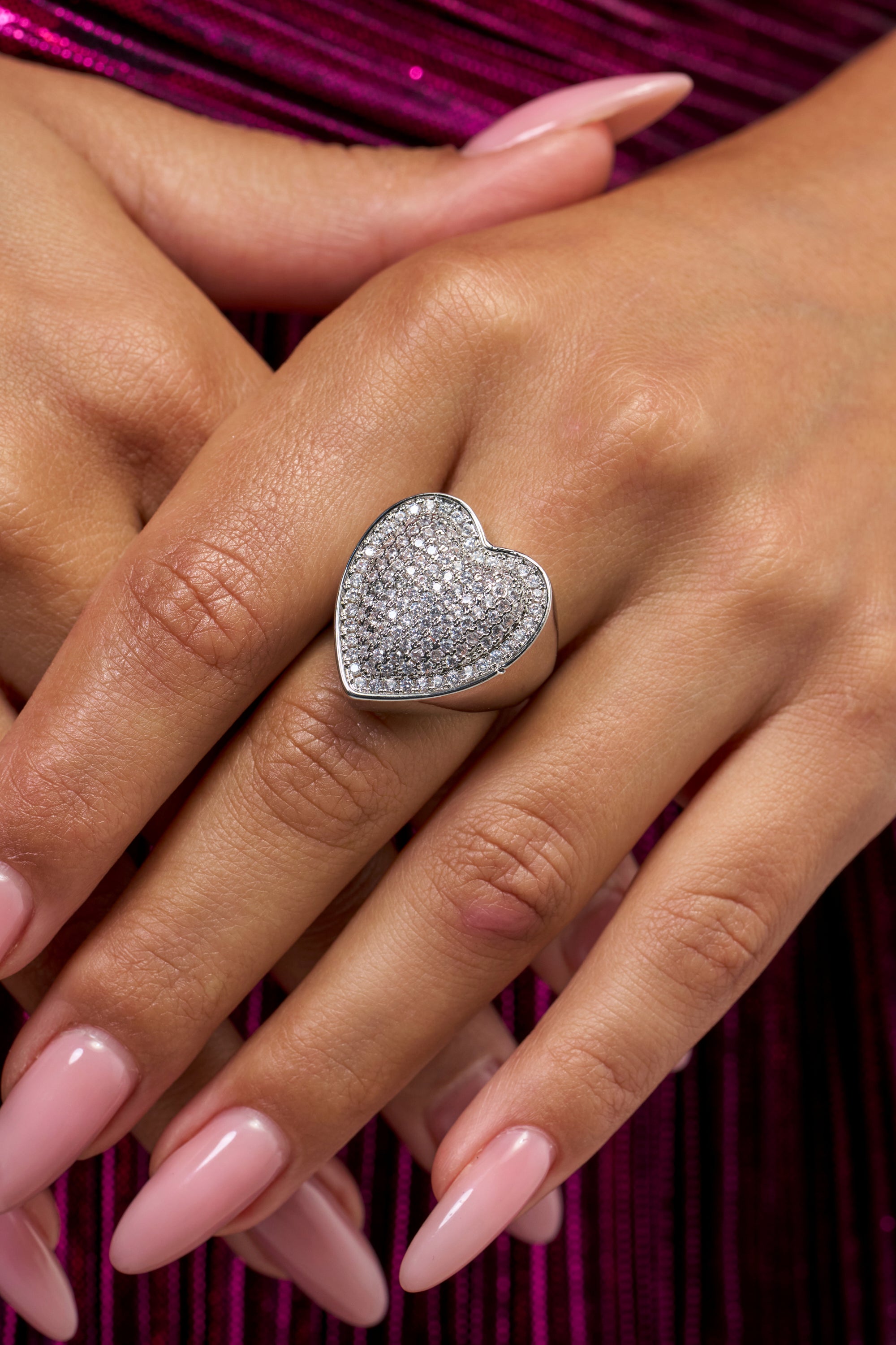 A close-up of hands with light pink manicured nails wearing the LOVE U RING—a chunky heart ring encrusted with small, sparkling stones—set against a magenta, pleated fabric background, perfect for showcasing glamorous silver jewelry.