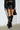 A person walking away, wearing a black pinstripe jacket, a black leather mini skirt with red trim, and knee-high AZALEA WANG RHODES BLACK CROC WEDGE BOOTS, while holding a small black handbag. The setting is a concrete floor with a white brick wall.