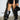 A person flaunts a black leather mini skirt with floral embroidery and a red trim, complemented by the stylish AZALEA WANG RHODES BLACK CROC WEDGE BOOT. They hold a chic black handbag, standing confidently against a light-colored brick wall.