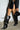 A person flaunts a black leather mini skirt with floral embroidery and a red trim, complemented by the stylish AZALEA WANG RHODES BLACK CROC WEDGE BOOT. They hold a chic black handbag, standing confidently against a light-colored brick wall.
