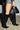 A person wearing the AZALEA WANG RHODES BLACK CROC WEDGE BOOT with knee-high design, pointed toes, and heels, alongside a black leather mini skirt, carries a black handbag. The background features a light-colored wall and floor.