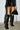 A person is seen from the back, wearing a short black leather skirt paired with the AZALEA WANG RHODES BLACK CROC WEDGE BOOT, which are knee-high and feature wedge stiletto heels. They're holding a faux crocodile leather handbag in their right hand. The backdrop is a white brick wall, with several black strings hanging from their outfit.