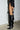 Against a white brick wall, an individual showcases an edgy ensemble featuring a black miniskirt with red trim and a black jacket. The bold look is completed with the AZALEA WANG RHODES BLACK CROC WEDGE BOOT, known for its knee-high faux crocodile leather design and pointed toes. Long black cords dangling from both the skirt and jacket enhance the overall stylish appearance.