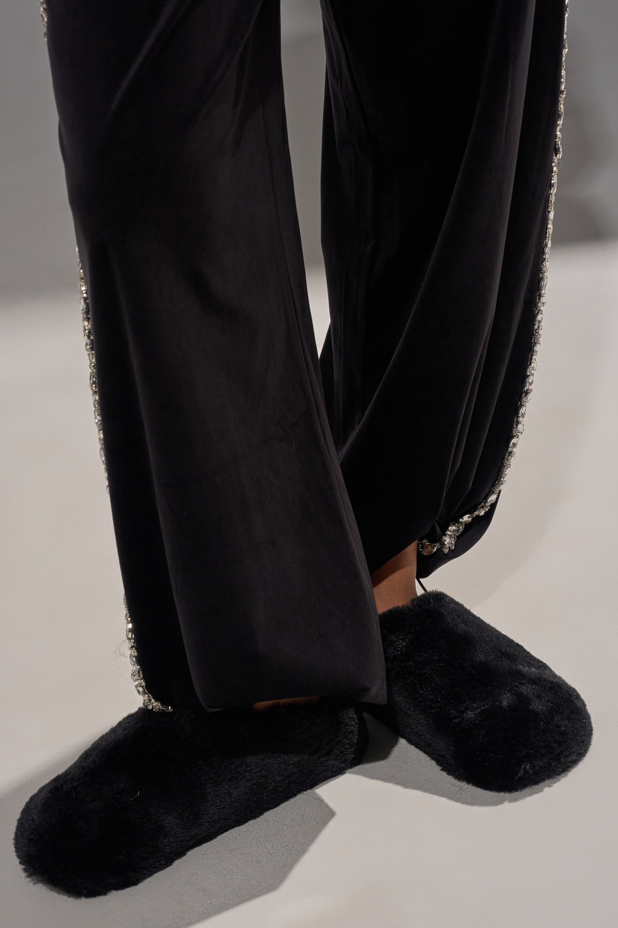 Wearing black wide-legged pants with beaded trim and the AKIRA RHYS BLACK FUZZY FLAT, a person stands on a light gray surface with legs crossed at the ankles.