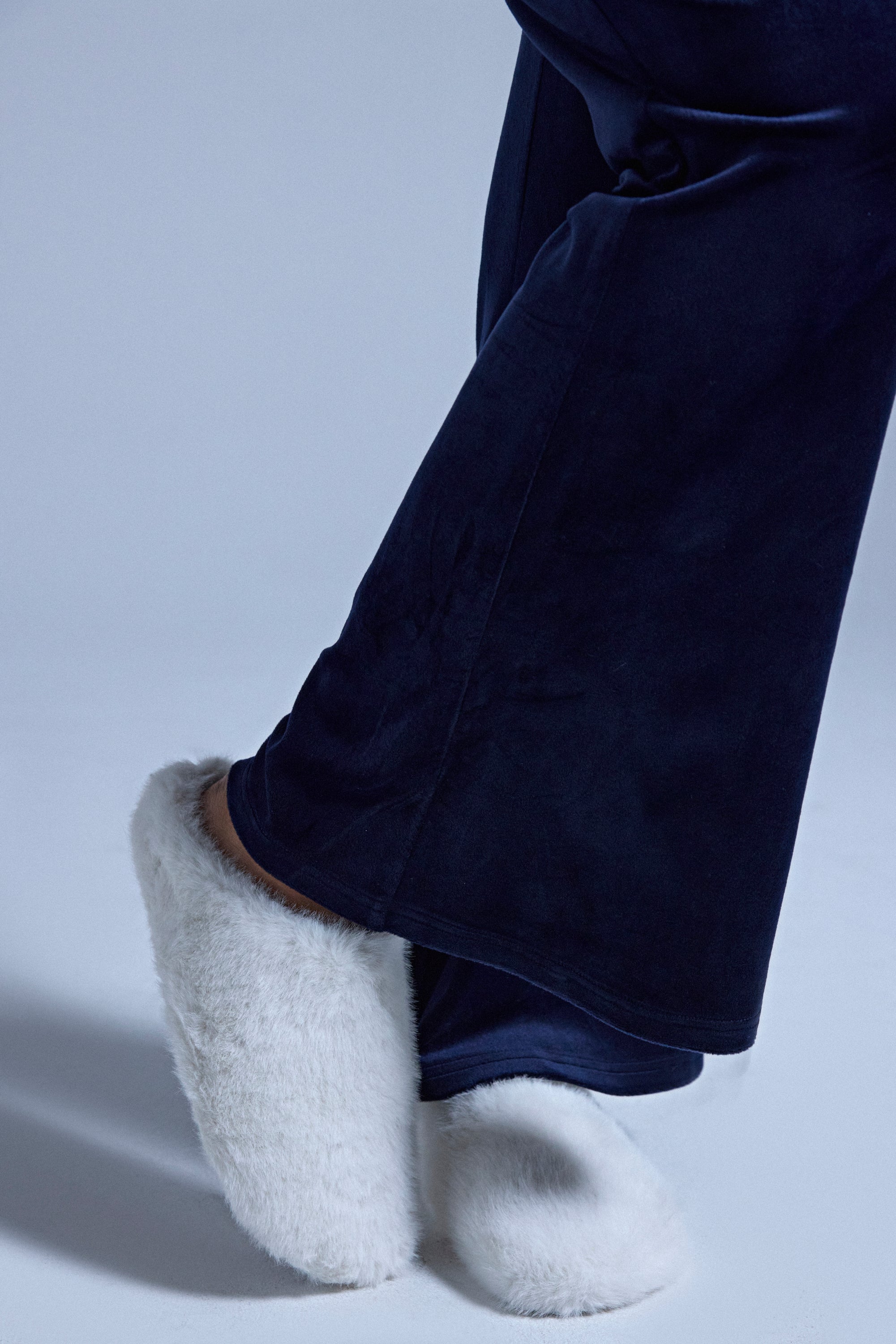 Someone stands on a light surface with one leg crossed, wearing dark blue pajama pants and the AKIRA RHYS IVORY FUZZY FLAT slippers featuring a cozy fleece-lined interior.