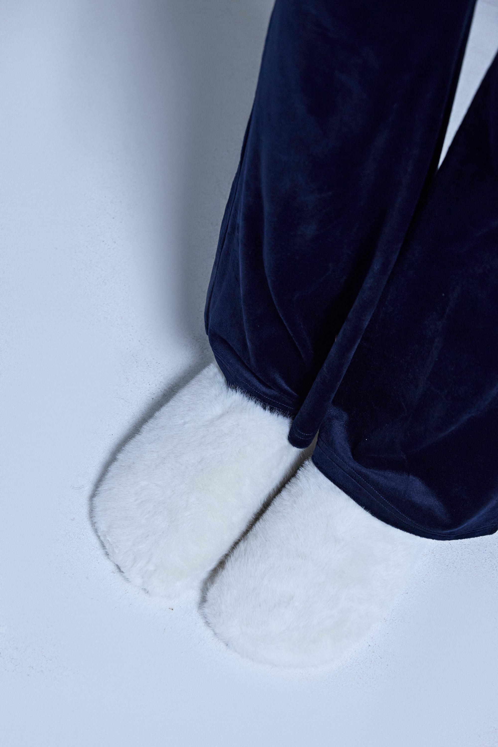 A person stands on a light floor, visible from the knees down, wearing dark blue pajama pants and AKIRA RHYS IVORY FUZZY FLAT slippers with fleece lining and flat rubber soles.